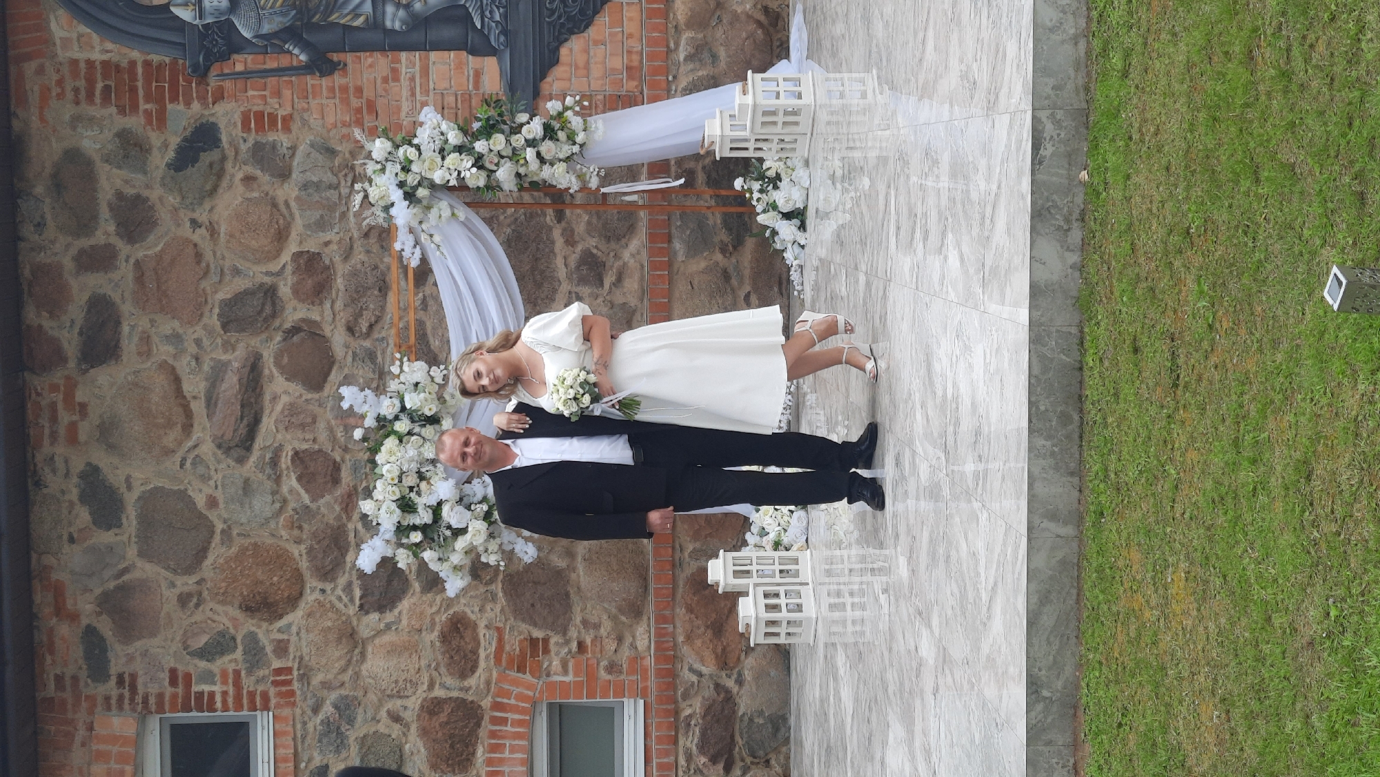A man and a woman are standing next to each other in front of a stone building. The man is wearing a black suit and a white shirt, and the woman is wearing a white dress. They are both smiling. There is a wedding arch behind them, decorated with white flowers and draped with white fabric. In front of them, there are two white lanterns. The ground is paved with light-colored tiles. To the left of the couple, there is a window in the stone wall. To the right, there is a mural of a knight in armor. The background is a stone wall with a brick facade on the left. The overall setting appears to be an outdoor wedding ceremony. The location is identified as Жодзішкаўскі сельскі Савет, Belarus.