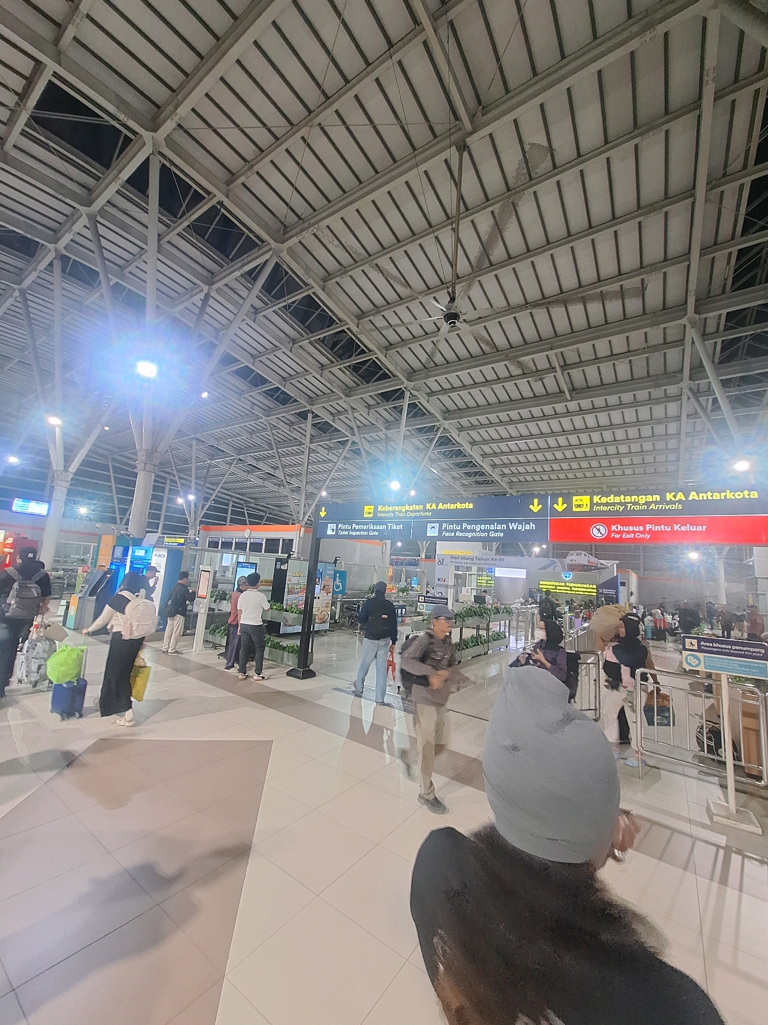 The image shows the interior of a train station, likely in Bekasi, Indonesia, given the Indonesian text on the signs. The station has a high, vaulted ceiling with exposed metal beams and a corrugated roof. Fluorescent lights are visible, some of which are bright enough to cause lens flare. Several signs with yellow and blue backgrounds are mounted on poles. One sign indicates "Keberangkatan KA Antarkota" (Intercity Train Departures) with an arrow pointing left and another sign says "Kedatangan KA Antarkota" (Intercity Train Arrivals) with an arrow pointing right. Another sign reads "Pintu Pengenalan Wajah" (Face Recognition Gate) and "Khusus Pintu Keluar" (For Exit Only).
The floor is tiled in a light color with some darker rectangular sections. There are many people in the station, some are walking with luggage, others are standing in queues or near information kiosks. A woman in the foreground is seen from behind, wearing a gray head covering and a dark jacket, holding a mobile phone. To her left, a woman with a large backpack and a green bag on wheels is walking. To the right, a group of people are gathered near a barrier. Various pieces of luggage, including suitcases and backpacks, are visible throughout the scene. The overall impression is of a busy transit hub.