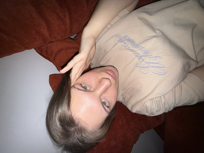 A woman lies on an orange sofa, her head resting on a cushion. She wears a beige t-shirt with a light blue cursive design. Her hair is parted to the side, and her hand is near her forehead. She gazes directly at the camera with green eyes. The background behind the sofa is a plain white wall. The angle is slightly tilted.