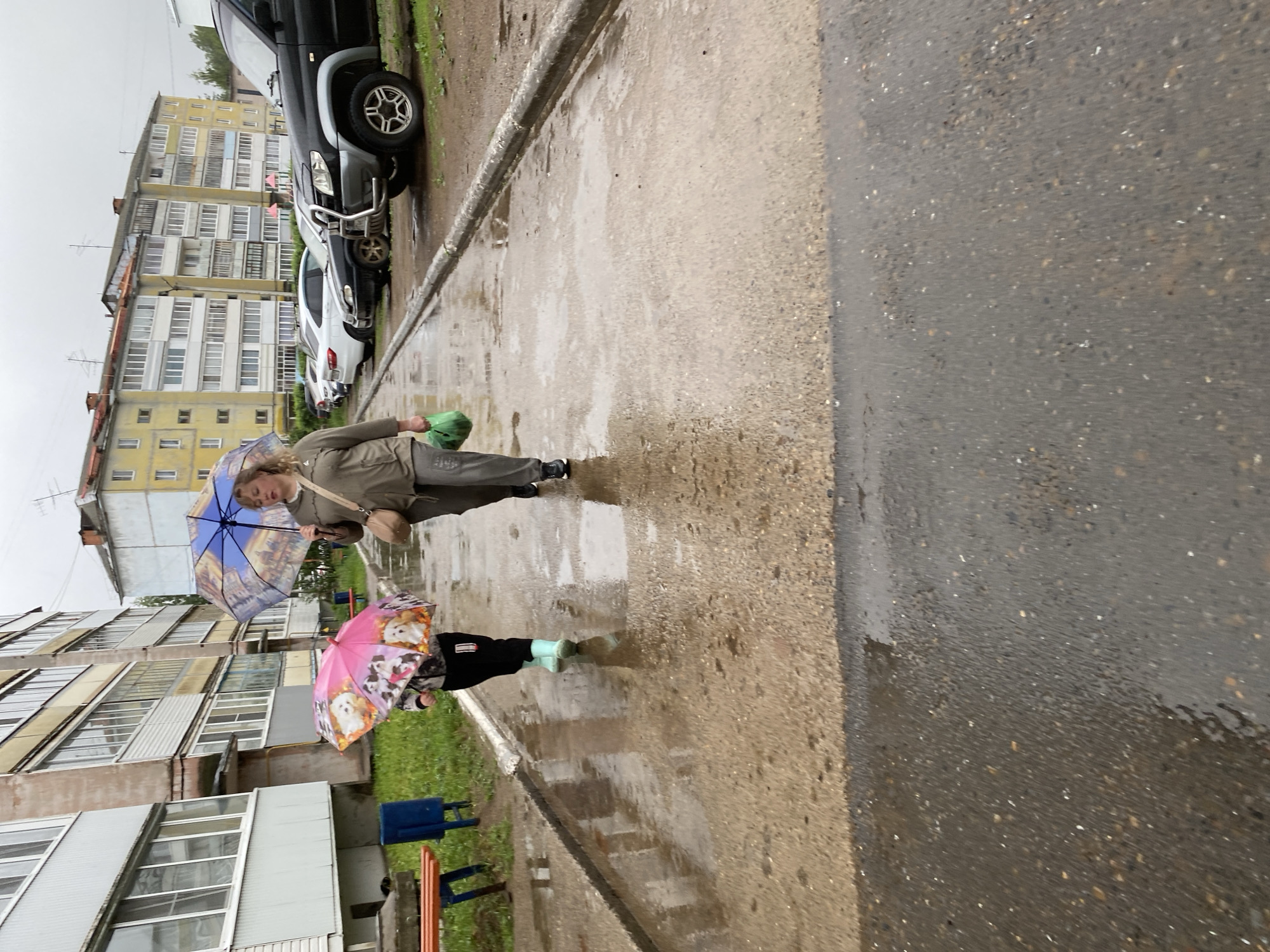 A street scene in Noviy Urengoy, Russia, shows an adult and a child walking in the rain, each carrying an umbrella. The adult, dressed in neutral-toned clothing, carries a blue umbrella with architectural depictions, and a green bag. The child, wearing black pants and light-colored boots, holds a pink umbrella adorned with puppies. The buildings are multi-story, Soviet-era apartment blocks. Parked cars line one side of the street. The road and sidewalk are wet, suggesting recent or ongoing rainfall. The sky is overcast, further indicating rainy weather. The benches from a playground are visible near the child.