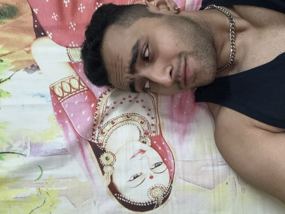 A man with dark hair and a silver chain necklace lies on a bed with a colorful print. The print depicts a woman with ornate jewelry and traditional clothing, likely an Indian or South Asian cultural representation. The man's expression is somewhat quizzical or skeptical. He wears a black tank top, and his arm is visible in the lower right corner. The background is slightly out of focus.