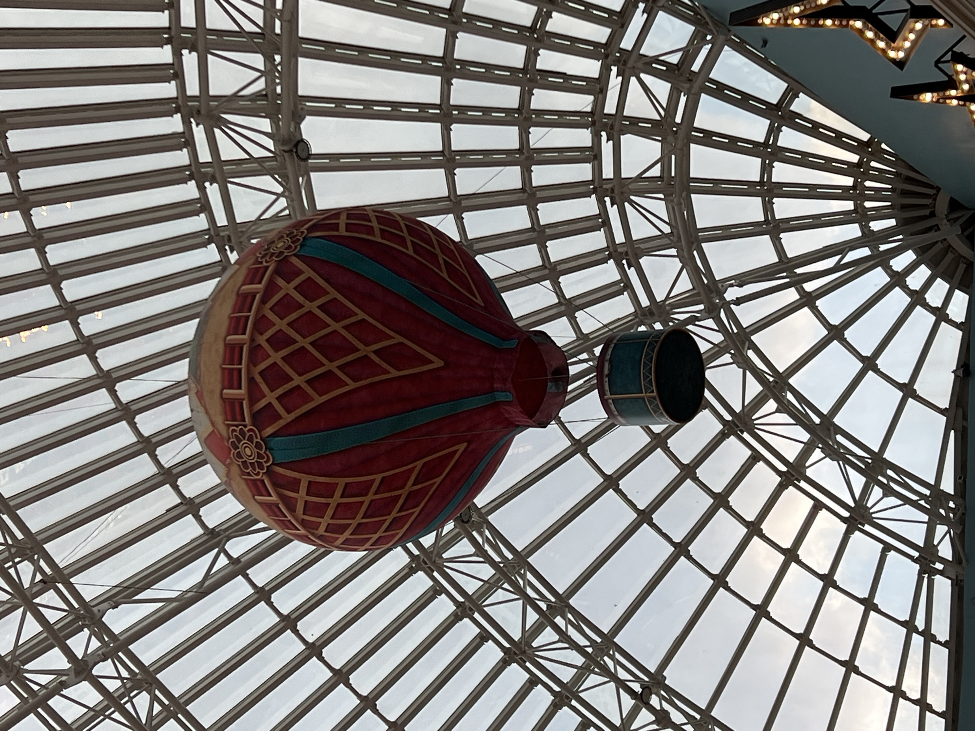 This is a low-angle, eye-level shot looking up at a decorative hot air balloon suspended from the ceiling of a large, indoor structure. The ceiling is a geodesic dome made of white metal beams and clear glass panels, allowing natural light to filter through. The hot air balloon is red with a pattern of yellow diamonds and turquoise stripes. A smaller, cylindrical object, possibly a basket or gondola, is attached to the balloon. The balloon appears to be a static decoration, not in flight. In the upper right corner of the frame, a section of a blue ceiling is visible, adorned with decorative star-shaped lights. The lighting suggests it could be daytime, given the natural light coming through the dome, but the overall ambiance is indoor and perhaps commercial, like a mall or amusement park. There are no people visible in the image. The scene is peaceful and the decorative elements suggest a whimsical or festive atmosphere. No visible text is present in the image.