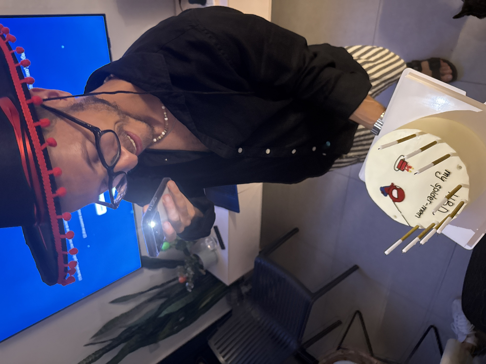 The image depicts a man wearing a sombrero and glasses, holding a smartphone to take a picture of a cake. The cake is decorated with a Spider-Man image and the words "Happy Birthday my spider-man". The man is wearing a black shirt and striped pants. The scene appears to be indoors, possibly a party setting, with a television screen in the background displaying a bright blue image. A chair and a plant are visible to the left. The man's attire and the cake suggest a birthday celebration. The image was captured in Tel-Aviv, Israel.
