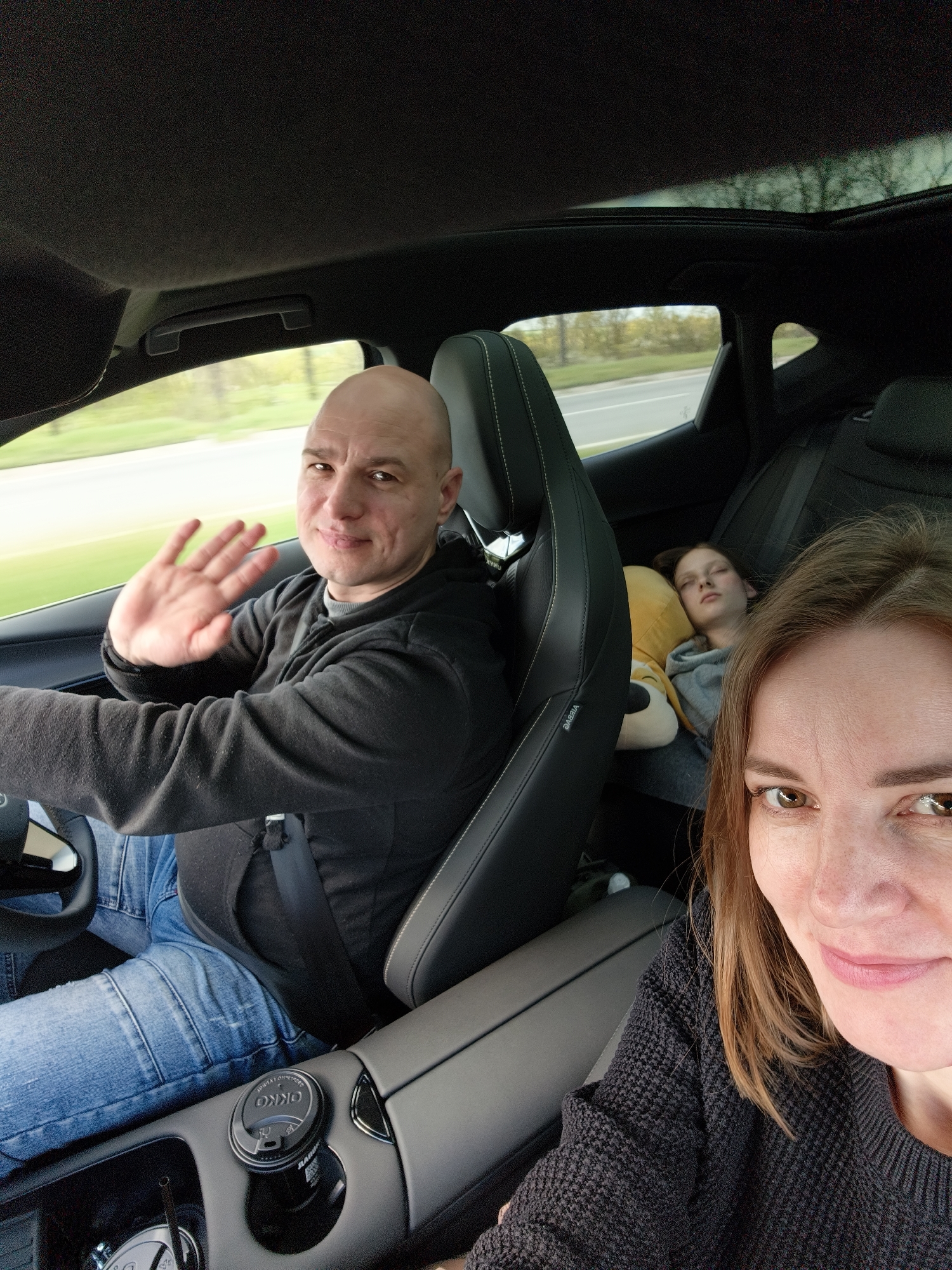 A man is driving a car, waving his hand. A woman is in the passenger seat, looking at the camera. A child is asleep in the back seat, resting against a yellow pillow. The man is bald, wearing a black sweater and blue jeans. The woman has brown hair and is wearing a black sweater. There is a coffee cup in the car's center console. The car has a sunroof, and trees are visible outside the windows. The location is somewhere near Krynychanska aglomeration, Ukraine.