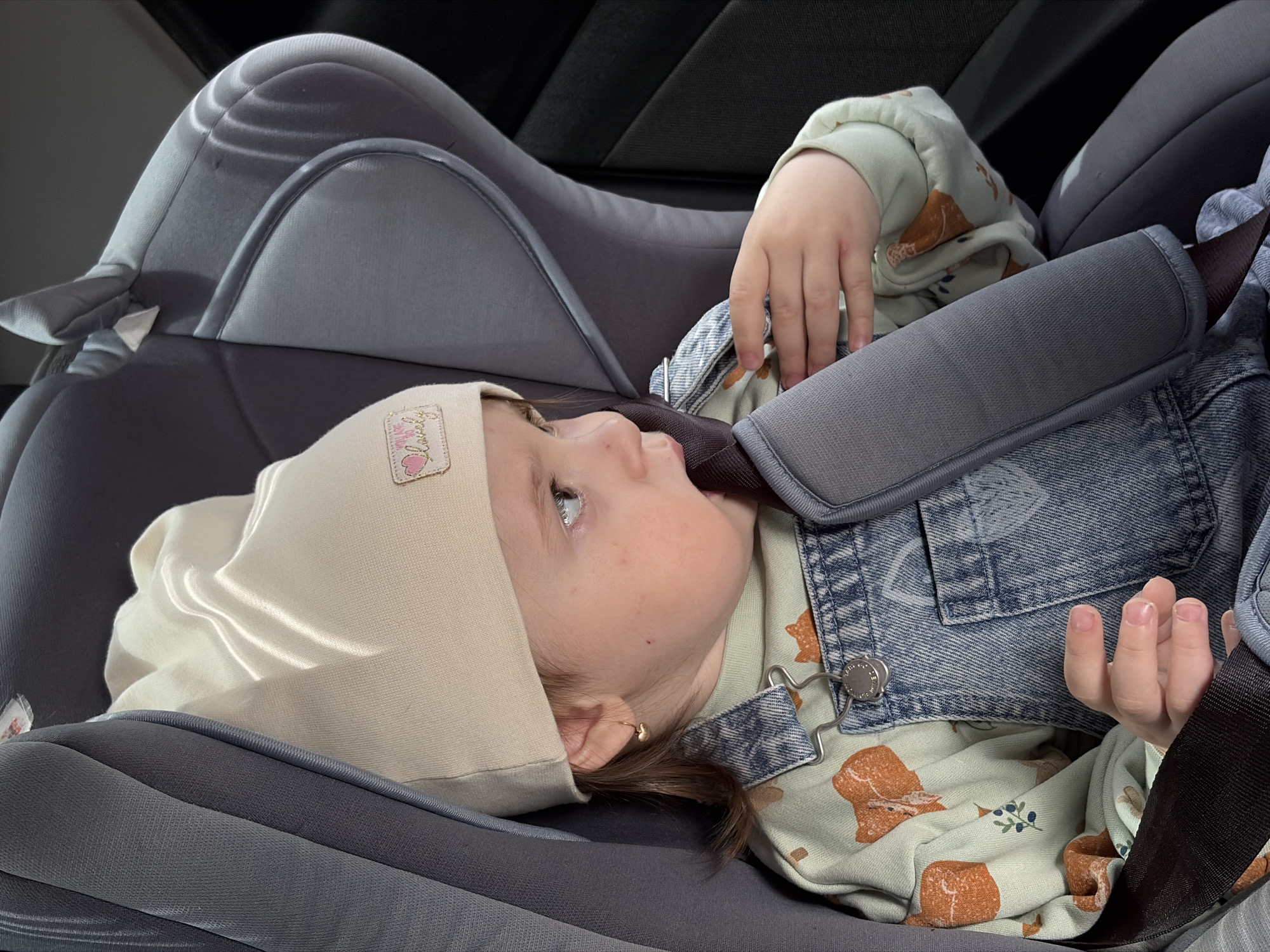 A young child, likely under the age of two, is secured in a grey car seat with a beige cap. The child is wearing a light green long-sleeved shirt with a pattern of brown teddy bears and blue berries, paired with denim overalls. The car seat is placed in a vehicle with a dark interior. The location is likely Chaikovsky, Russia.