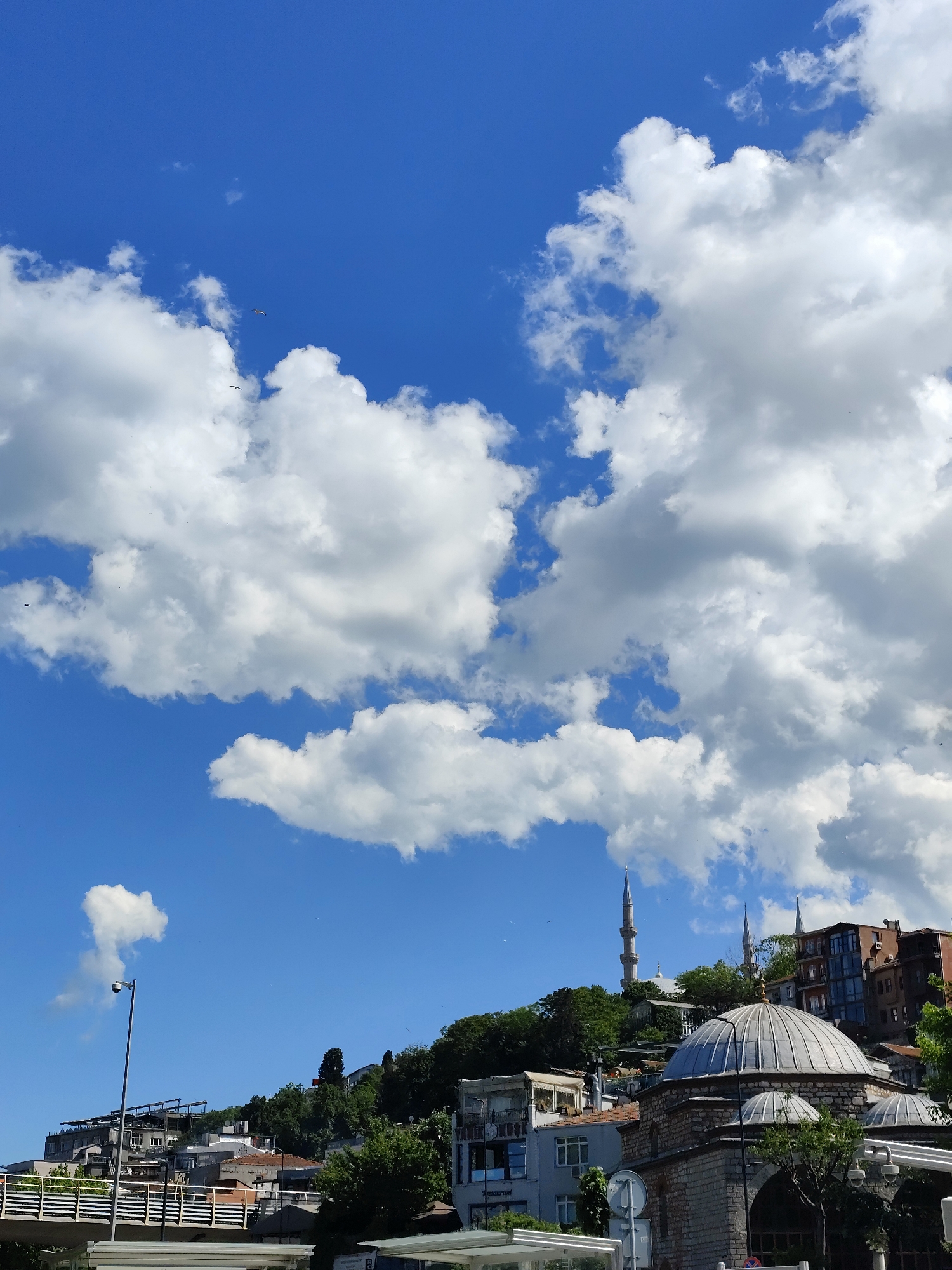 The image captures a view of Istanbul, Turkey, featuring a mix of architectural and natural elements under a partly cloudy sky. In the foreground, the domed roofs of a historical structure are visible, constructed from light-colored stone or brick. Behind this structure, the built environment rises up a hillside, including residential buildings and other structures. The skyline is punctuated by the minarets of a mosque, rising above the other buildings. The sky is a bright blue, with a significant portion filled with white cumulus clouds. Scattered birds are also visible in the sky. A street light is present on the left of the image. The scene is well-lit, suggesting it is daytime.