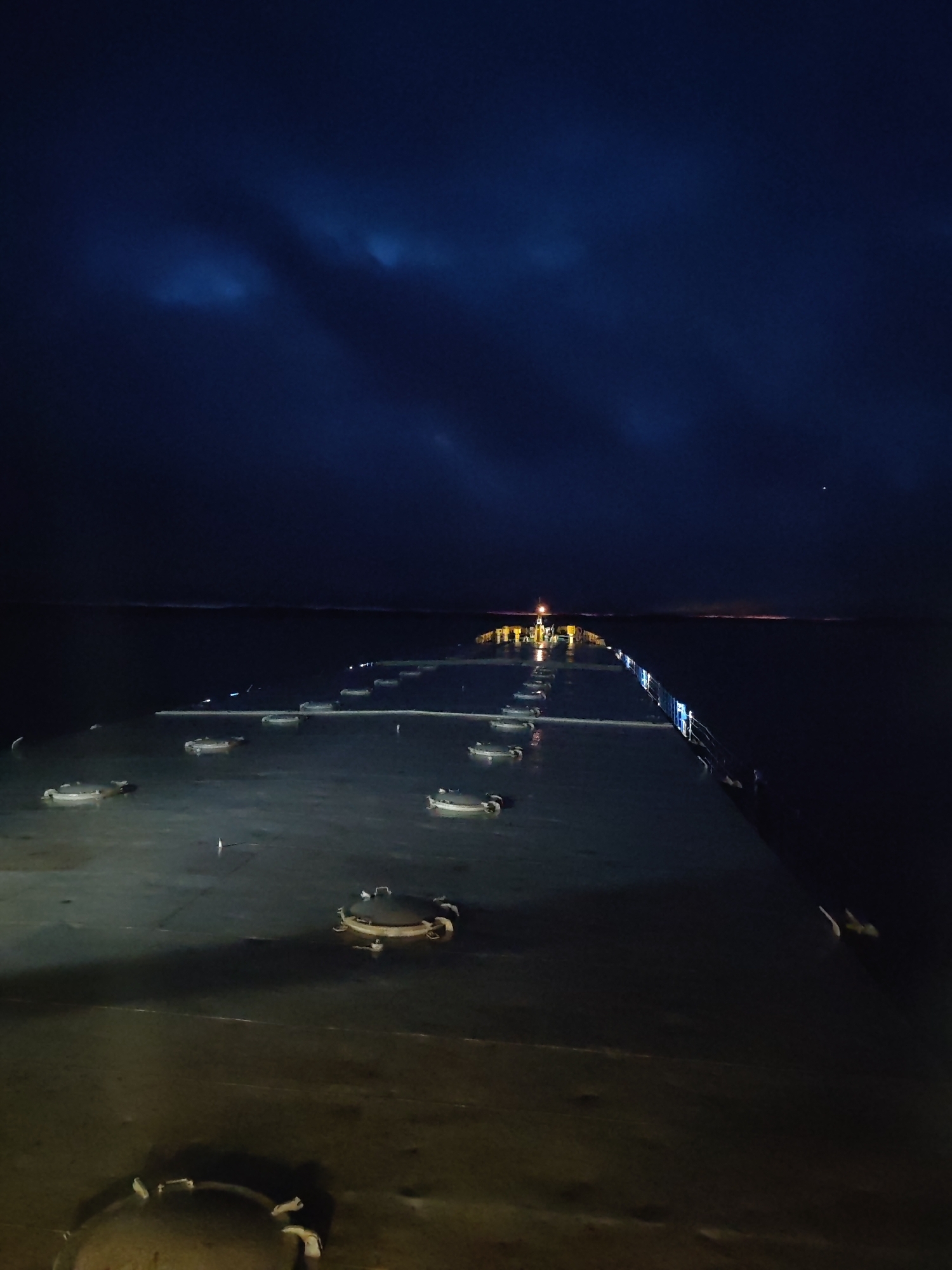 The image captures a nighttime scene from the deck of a large vessel, likely a ship or ferry, looking forward across its bow towards the horizon. The setting is at sea, under a dark, star-speckled sky with subtle, darker cloud formations visible, particularly towards the upper left.
The primary subject is the expansive deck of the vessel, which appears damp or reflective due to the ambient light. Numerous circular, raised hatch covers or manholes are distributed across the deck surface. A railing, consisting of vertical stanchions and horizontal bars, extends along the right side of the deck into the distance, showing faint reflections or lights.
In the mid-ground, a prominent, yellowish-orange light source, likely a mast light or navigational light of the vessel, illuminates a small section of the deck and structures around it. Beyond the vessel, the dark, open water stretches towards the horizon. A faint, reddish-orange glow is discernible on the far horizon to the right, possibly indicating distant land or another vessel. High in the dark sky, to the right of the central axis, a single, bright white star or planet is visible.
No people are present, and no specific activity or event is taking place, suggesting a moment of quiet transit at night. There is no visible text in the image. The overall mood is serene and contemplative, emphasizing the vastness of the sea and sky against the illuminated structure of the ship.