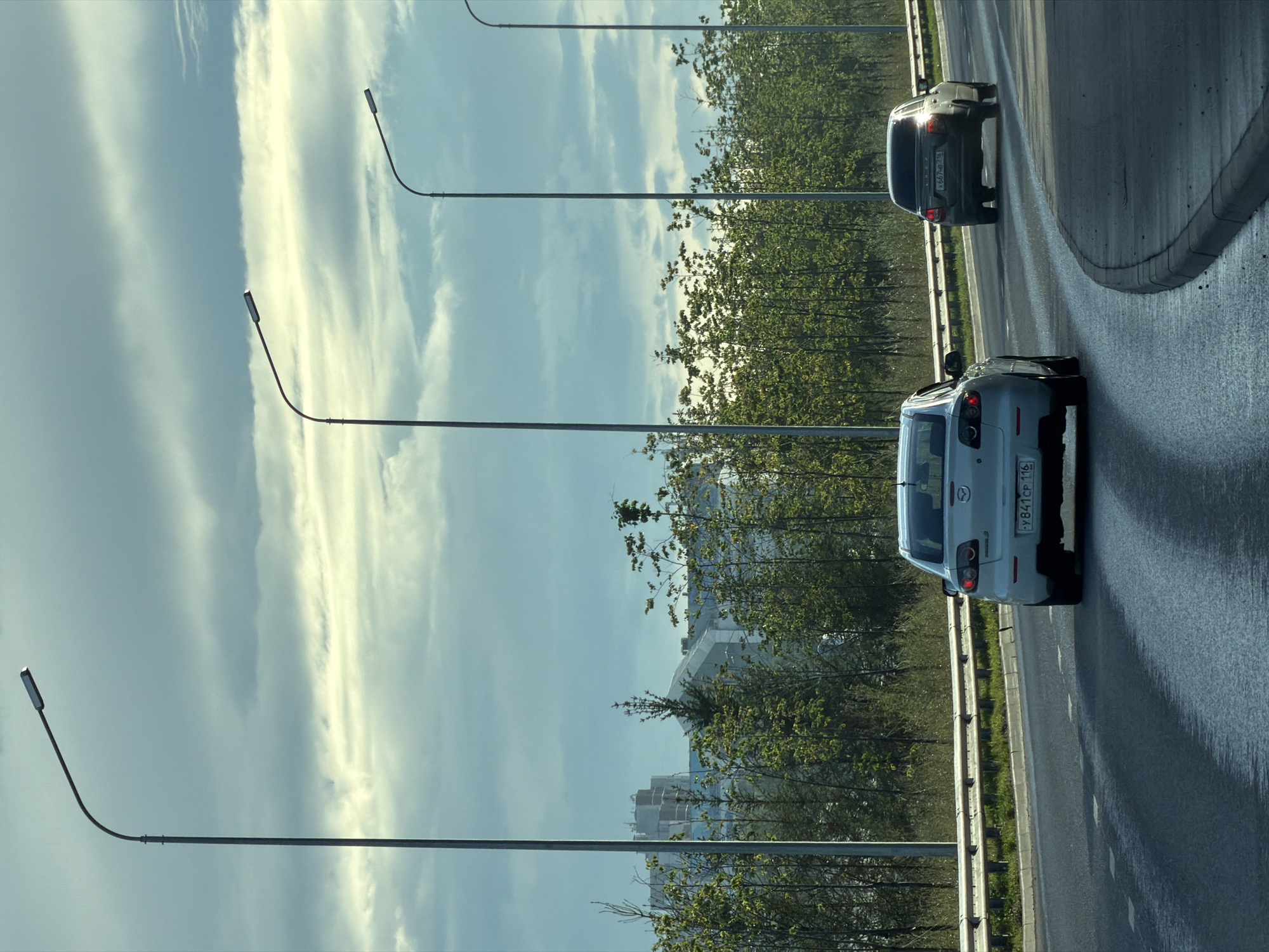 The image depicts a highway scene in Kazan, Russia, under a cloudy sky. Two cars are visible on the road: a light-colored Mazda in the foreground and a darker car further ahead. Tall streetlights line the side of the road, and a row of trees separates the highway from buildings in the distance. The road surface appears wet, possibly from recent rain, and reflects the overhead sky and streetlights. The overall atmosphere is overcast, suggesting early morning or late afternoon lighting.