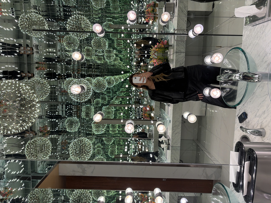 A woman in a black outfit holds a phone in a bathroom with mirrors creating an infinity effect. The room is illuminated by numerous spherical light fixtures, adding to the illusion of endless space. Modern, clear sinks with chrome fixtures sit on a white marble countertop. Details like tissue dispensers and a small black item are also visible on the counter.