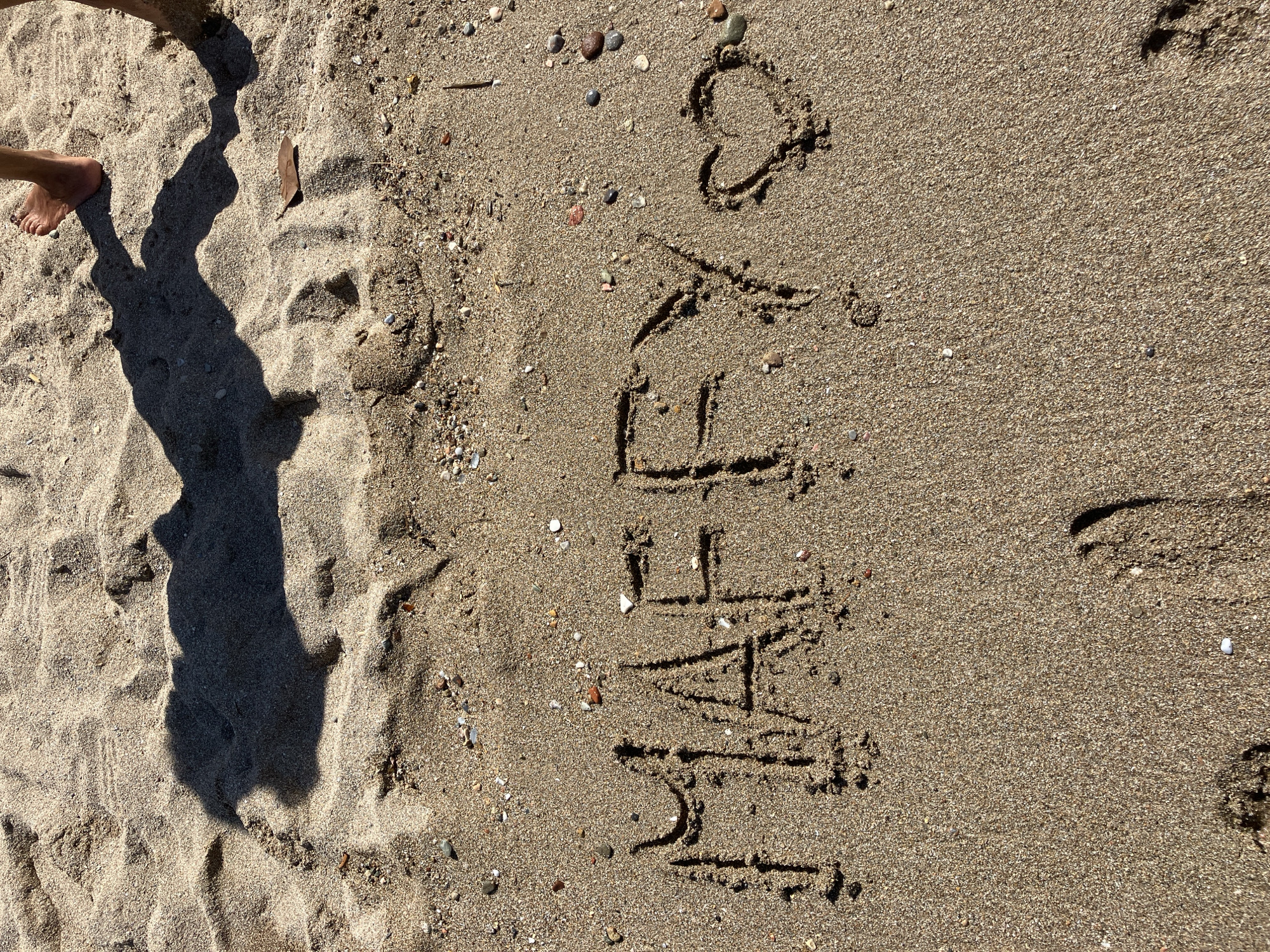 A beach scene in Paleo Faliro, Greece, features the word "MAFFY" written in the sand, followed by a heart symbol. The sand is composed of fine grains with scattered pebbles and shells. In the background, a shadow of two people is cast upon the rippled sand. One person's foot and ankle are visible at the top right of the frame, with the toes pointing towards the left. The overall lighting suggests a sunny day.