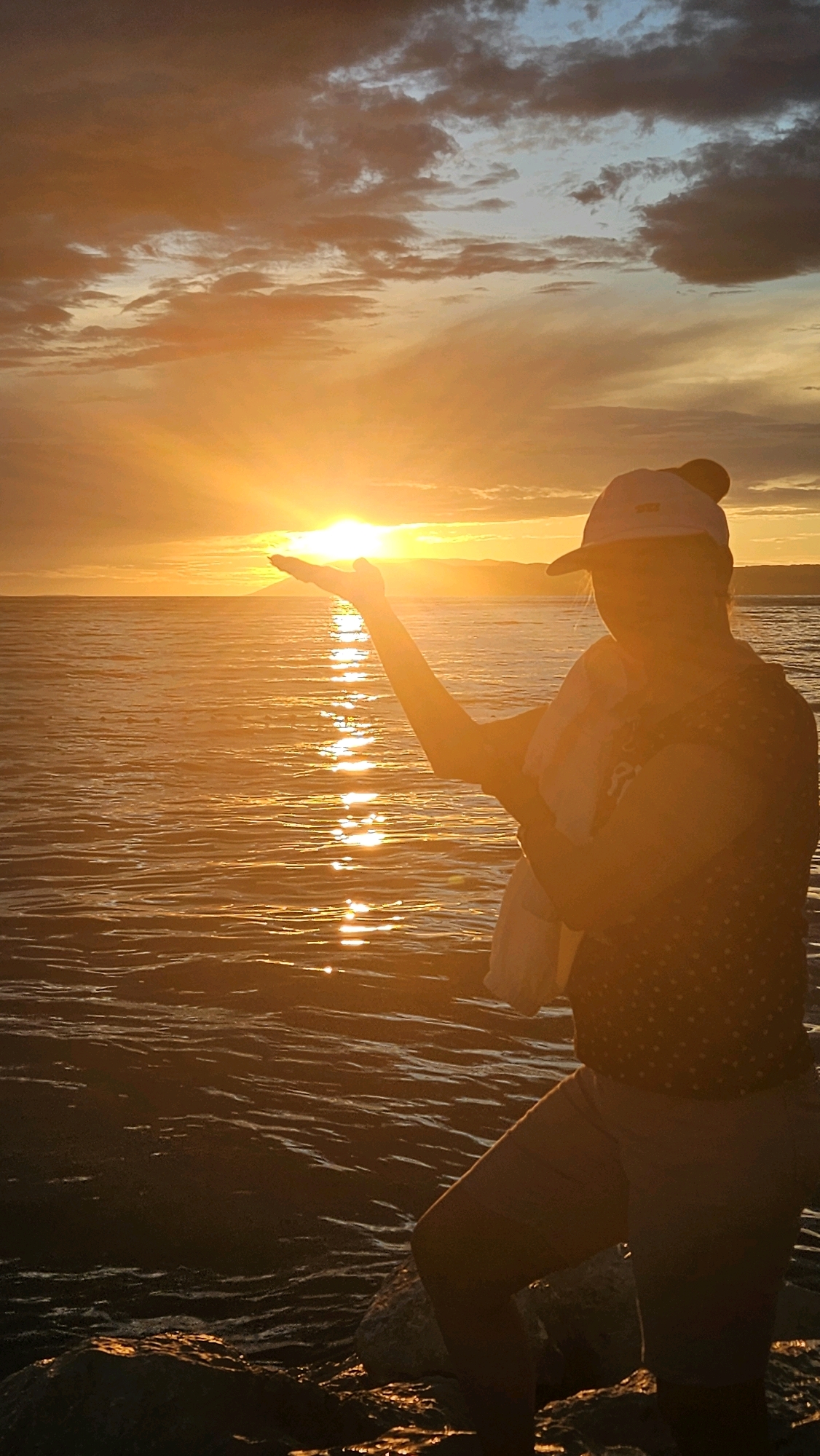 An over-the-shoulder, vertical, medium shot captures a person standing on a rocky shoreline at sunset, with their right arm extended and hand cupped, appearing to hold the setting sun. The sun, a bright orb of golden light, is positioned just above the horizon over a body of water. The sky is filled with dramatic, colorful clouds in shades of orange, yellow, and gray, with hints of blue. The water reflects the warm hues of the sunset, creating a shimmering path of light. The person is wearing a baseball cap and a patterned shirt, with a white towel draped over their shoulder. The rocky shore is dark and wet, suggesting it is at the edge of the water. The scene suggests a serene and picturesque moment at the end of the day.