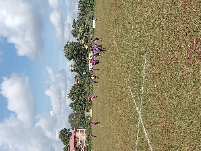 The image displays a grassy sports field in Meru, Kenya, under a blue sky with scattered cumulus clouds. White line markings are visible on the field. Approximately 18 to 20 individuals, appearing to be young adults or teenagers, are present. Most are attired in athletic wear, predominantly pink/magenta and purple jerseys, some with shorts or track pants. A concentration of 8-10 individuals is located in the mid-right portion of the field, suggesting a group activity or instruction. Other individuals are standing dispersed across the field. In the background, a multi-story building with a red roof is situated to the far left. Trees line the field's periphery, behind which a tall metal communication tower is visible, along with additional distant structures. Advertising banners, one yellow and black, are positioned near the background tree line.