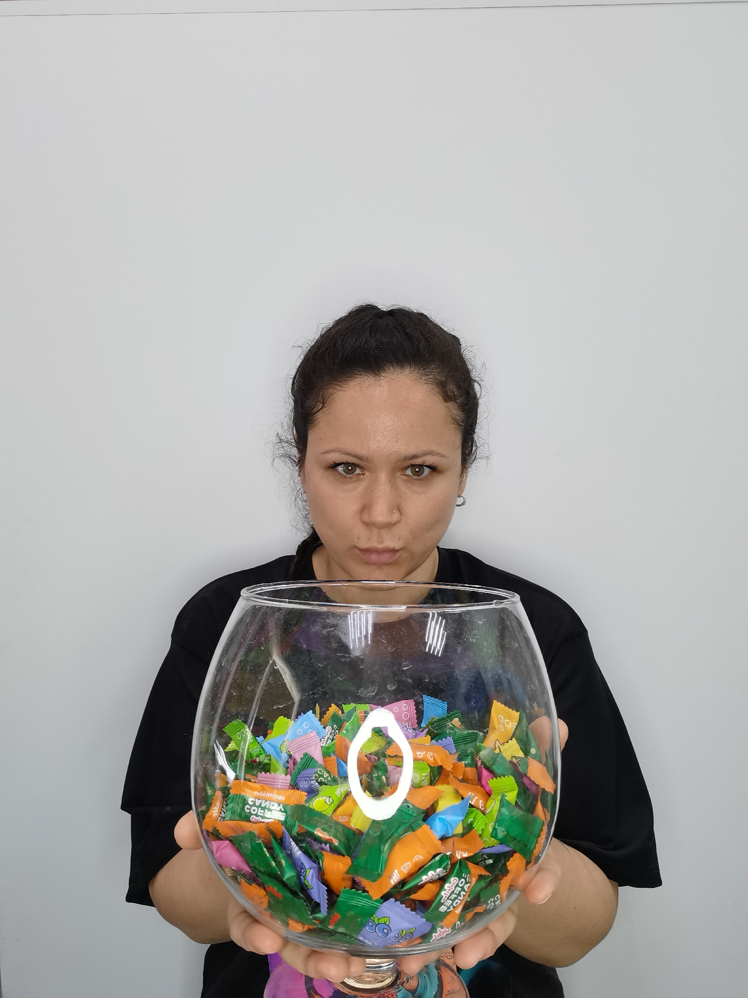 A woman with dark hair pulled back and wearing a black t-shirt holds a large glass bowl filled with wrapped candies. The candies are various colors including orange, green, pink, and purple. The woman is looking directly at the camera with a slightly pursed expression. The background is a plain white wall. There is no discernible information in the image to confirm or deny a location in Chaikovsky, Russia.
