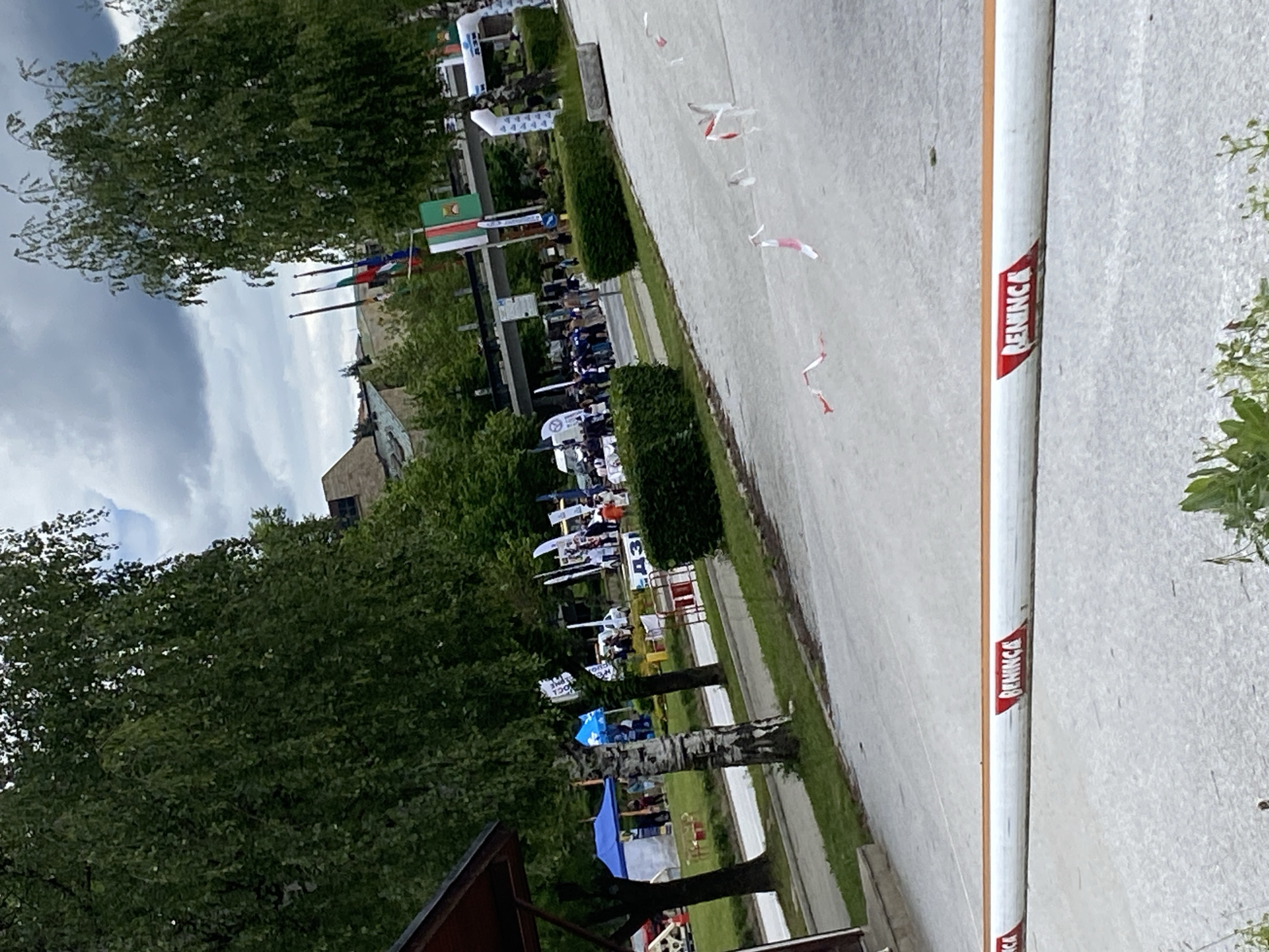 The image depicts an outdoor event, likely a race or a festival, in the city of Smolyan, Bulgaria. In the foreground, a white barrier with red markings, displaying the word "BENINCA," stretches across the frame. The background reveals a gathering of people, tents, flags, and trees. The environment appears to be a park or open area within the city, with buildings and a cloudy sky visible in the distance. The overall scene suggests a lively event, possibly a competition or a public celebration, taking place on a cloudy day.