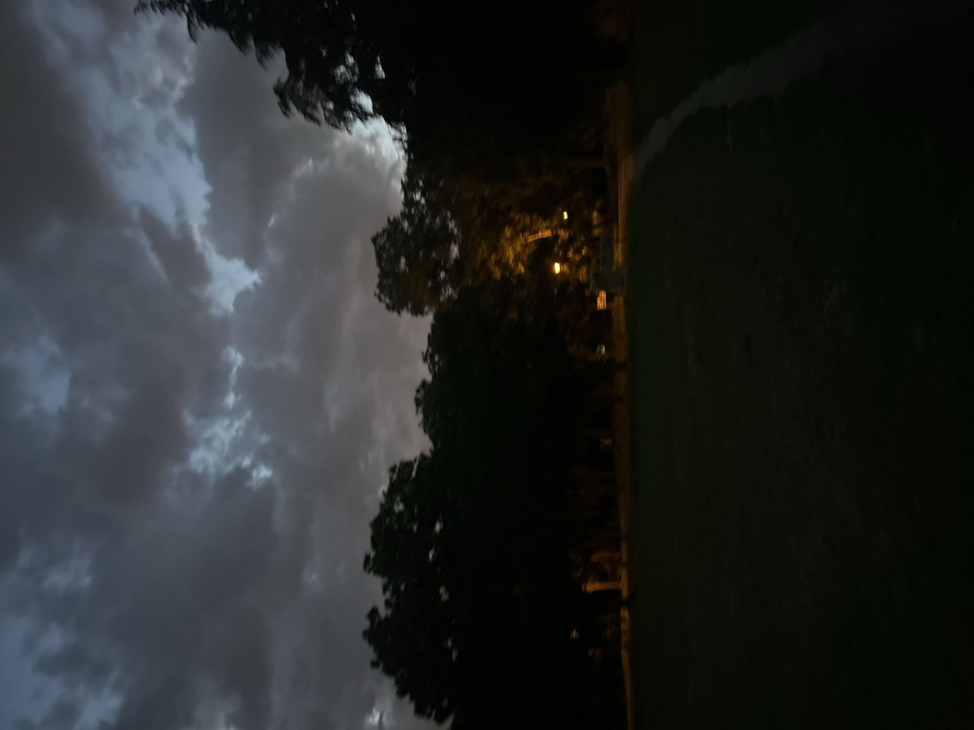 The image shows a park in Plovdiv, Bulgaria, at dusk or night. The sky is overcast with dark, heavy clouds filling the left side of the frame. A row of trees, silhouetted against the sky, separates the sky from the park. Streetlights, visible as yellow dots through the trees, illuminate a path running along the edge of a green field. The field takes up the right side of the image and appears dark due to low lighting. The overall scene conveys a sense of gloominess and stillness.