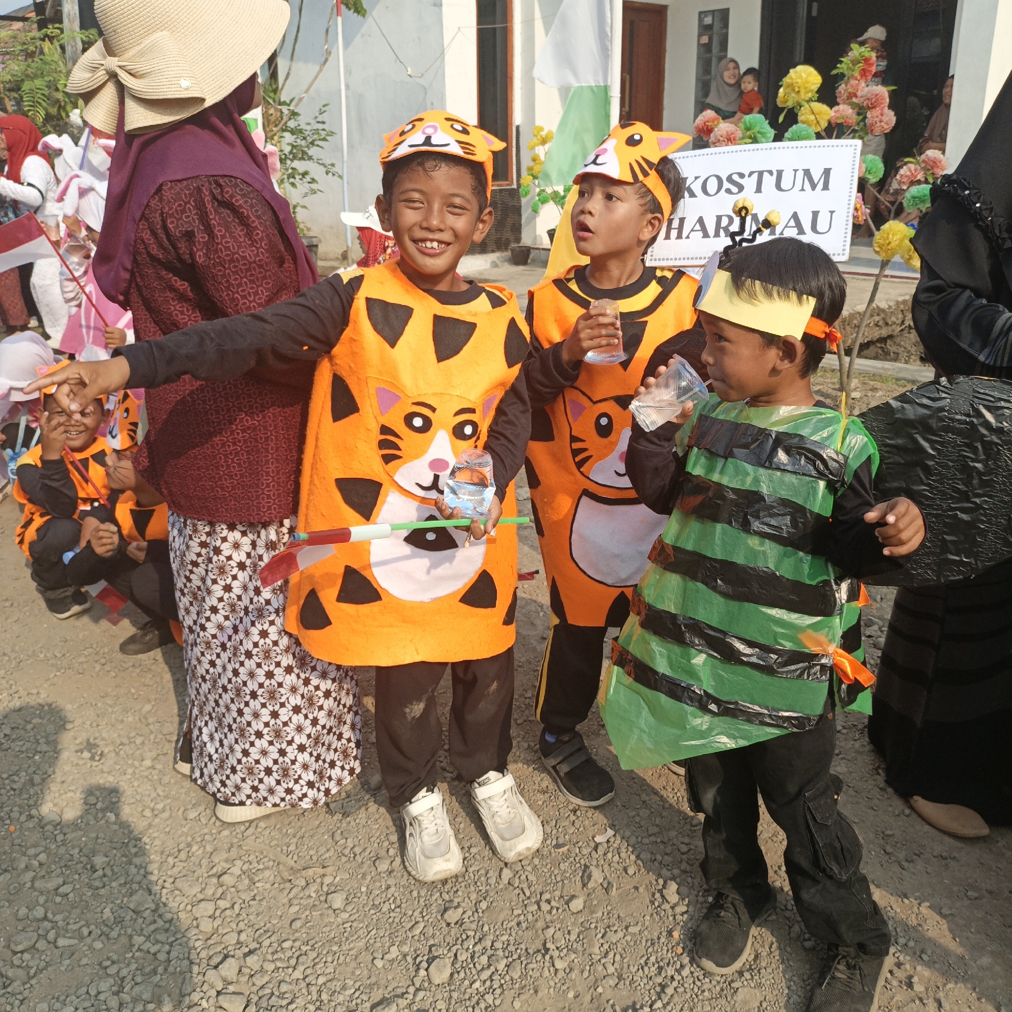 In Tanahbaya, Indonesia, children participate in a parade wearing costumes. The central figures are three boys dressed in animal-themed outfits. Two of the boys wear tiger costumes, complete with orange and black striped vests and matching headbands featuring tiger faces. One tiger-costumed boy holds a green and red striped wand and a plastic bottle of water. The third boy is dressed as a bee, with a green body adorned with black stripes, also wearing a yellow headband. Behind the children, a sign reads "KOSTUM HARI MAU," suggesting a costume day event. Other individuals, including adults, are visible in the background, some wearing traditional attire and holding flags. The ground is covered in gravel, indicating an outdoor setting, possibly a street or a field. The overall atmosphere suggests a community event or celebration.