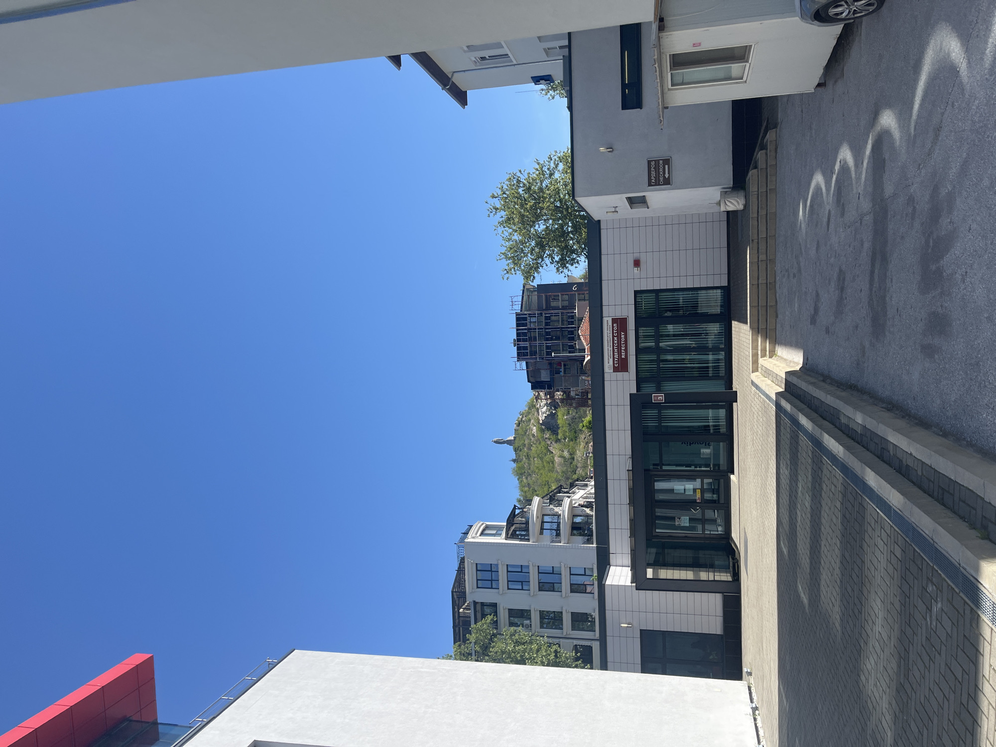 The image shows a view of the city of Plovdiv, Bulgaria, from an alleyway between modern buildings. The sky is clear blue. A construction site is visible in the background on a hill. A monument stands on a hill in the distance. An establishment with a sign in Bulgarian that translates to "Student Refectory" is visible. Signs for "Checkroom" can also be seen. A parked car is visible at the top right of the image. The alleyway is paved with bricks and concrete.