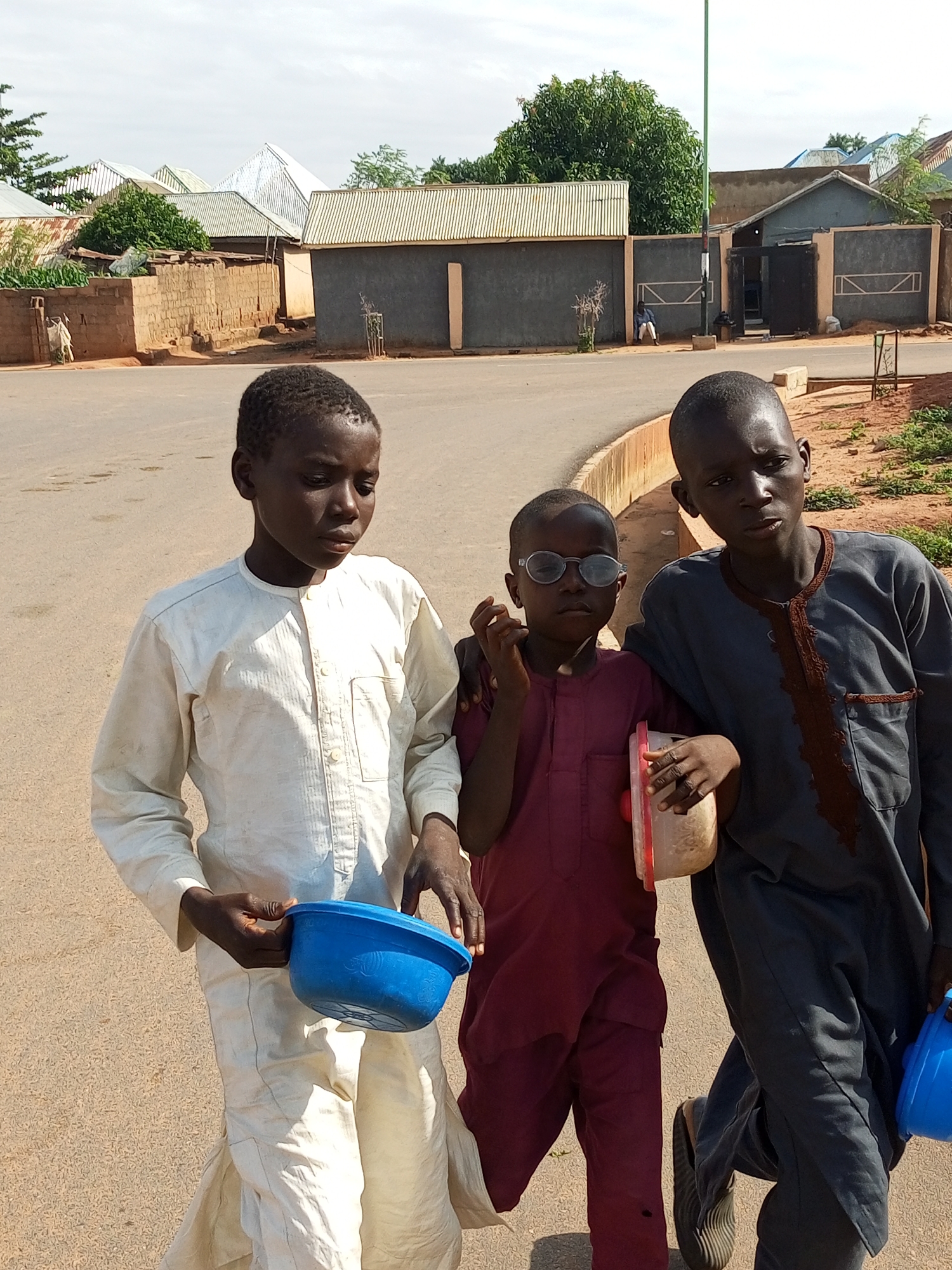 In Gombe, Nigeria, three young boys walk along a dirt road. The boy on the left wears a white tunic and holds a blue bowl. The middle boy, wearing a maroon outfit and round glasses, carries a clear plastic container. The boy on the right is dressed in a dark blue tunic. In the background, buildings with corrugated metal roofs and brick walls are visible, along with some greenery. The sky is overcast.