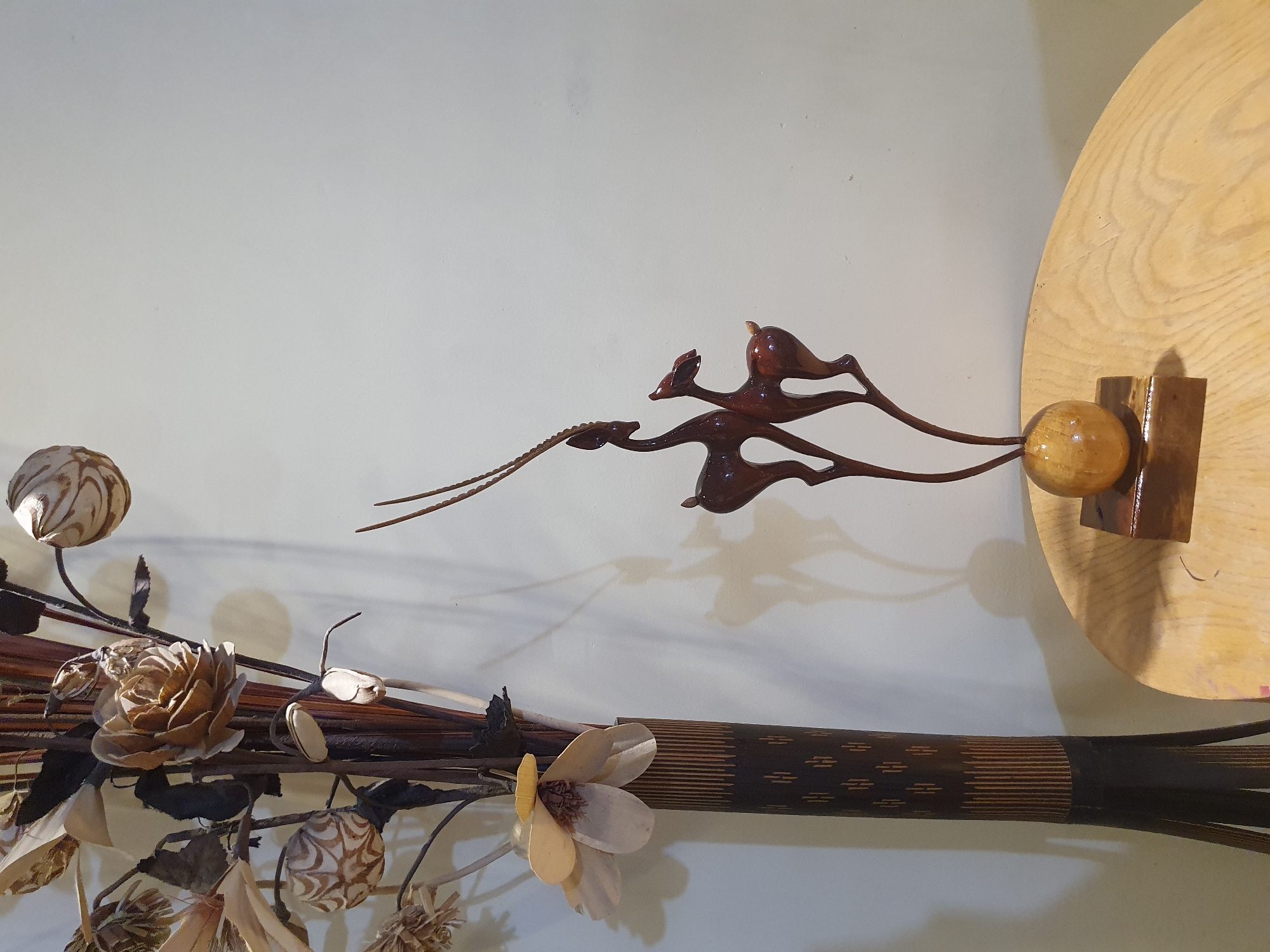 A decorative dried floral arrangement stands in a dark, patterned vase on the left side of the frame. To the right of the vase, a wooden sculpture of two gazelles is positioned on a light-colored, circular wooden surface. The gazelles are dark brown, with one slightly taller than the other. Behind the objects, a plain, off-white wall is visible, with shadows cast by the sculpture and vase. The scene is indoors, with no discernible city or specific location identified.