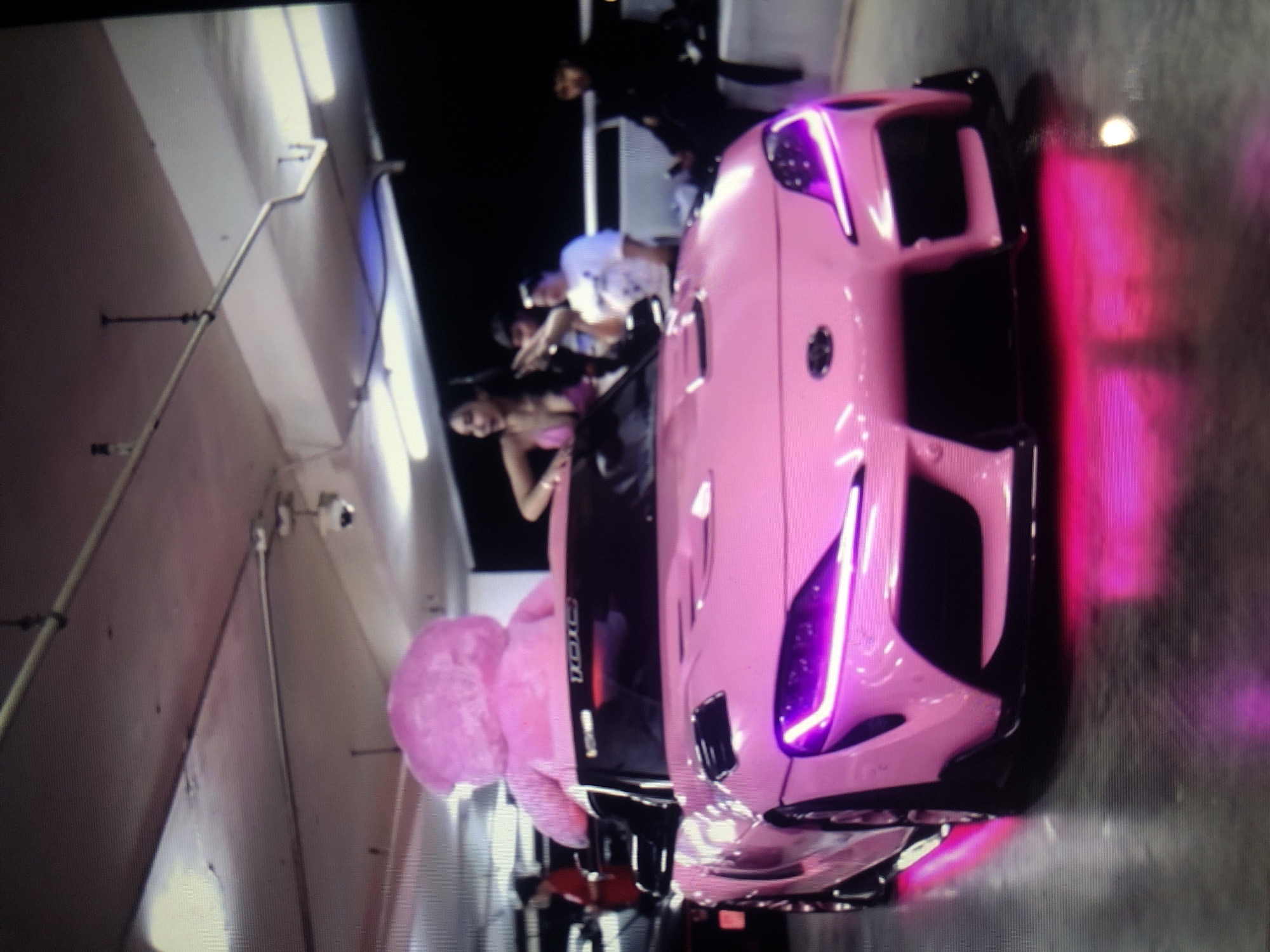 The image shows a pink Toyota Supra parked indoors, presumably in a parking garage. A large pink teddy bear is on the hood and roof of the car. People are gathered around the car, some standing and taking pictures. The car has pink underglow and its headlights are illuminated with purple lights. The setting appears to be a public parking garage, indicated by the ceiling fixtures and support beams visible. The image does not definitively state the location is Lhokseumawe, Indonesia.