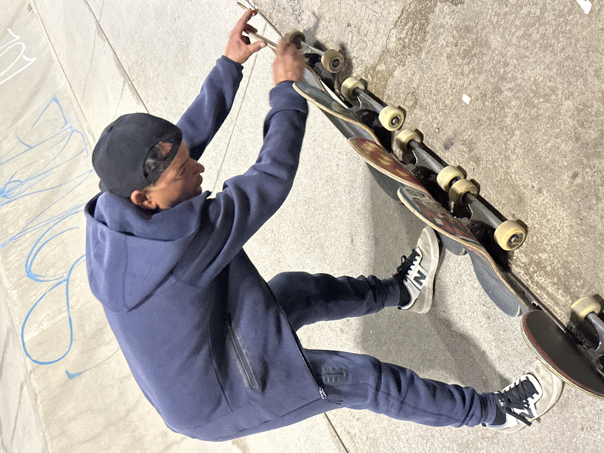 A man wearing a navy hooded sweatshirt and matching sweatpants and a black baseball cap is squatting beside a concrete wall, holding a pair of pliers and attending to a stack of three skateboards. The man is wearing New Balance shoes. The scene is outdoors, likely in an urban skatepark or similar location. There is graffiti on a nearby wall. This location cannot be determined to be in Barcelona, Spain from the image provided.