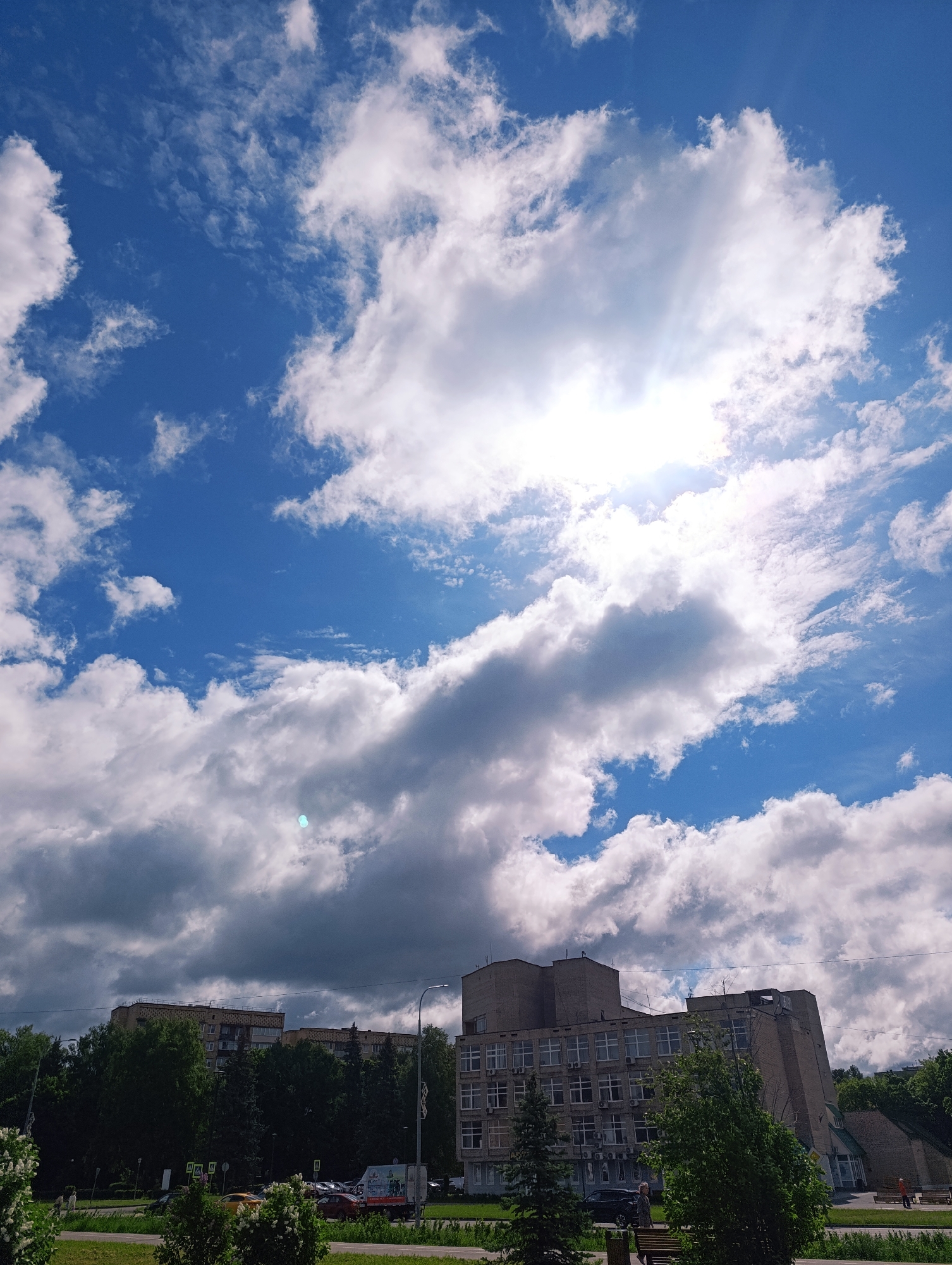 The image shows a building with a sky filled with clouds and sun in the city of Unknown, Unknown. The building is a multi-story structure with many windows. There are trees and vegetation around the building. In front of the building, there is a road and a park-like area with benches. Several people and vehicles are visible, including parked cars, a truck, and pedestrians. The sky is mostly blue with scattered white clouds and bright sunlight.