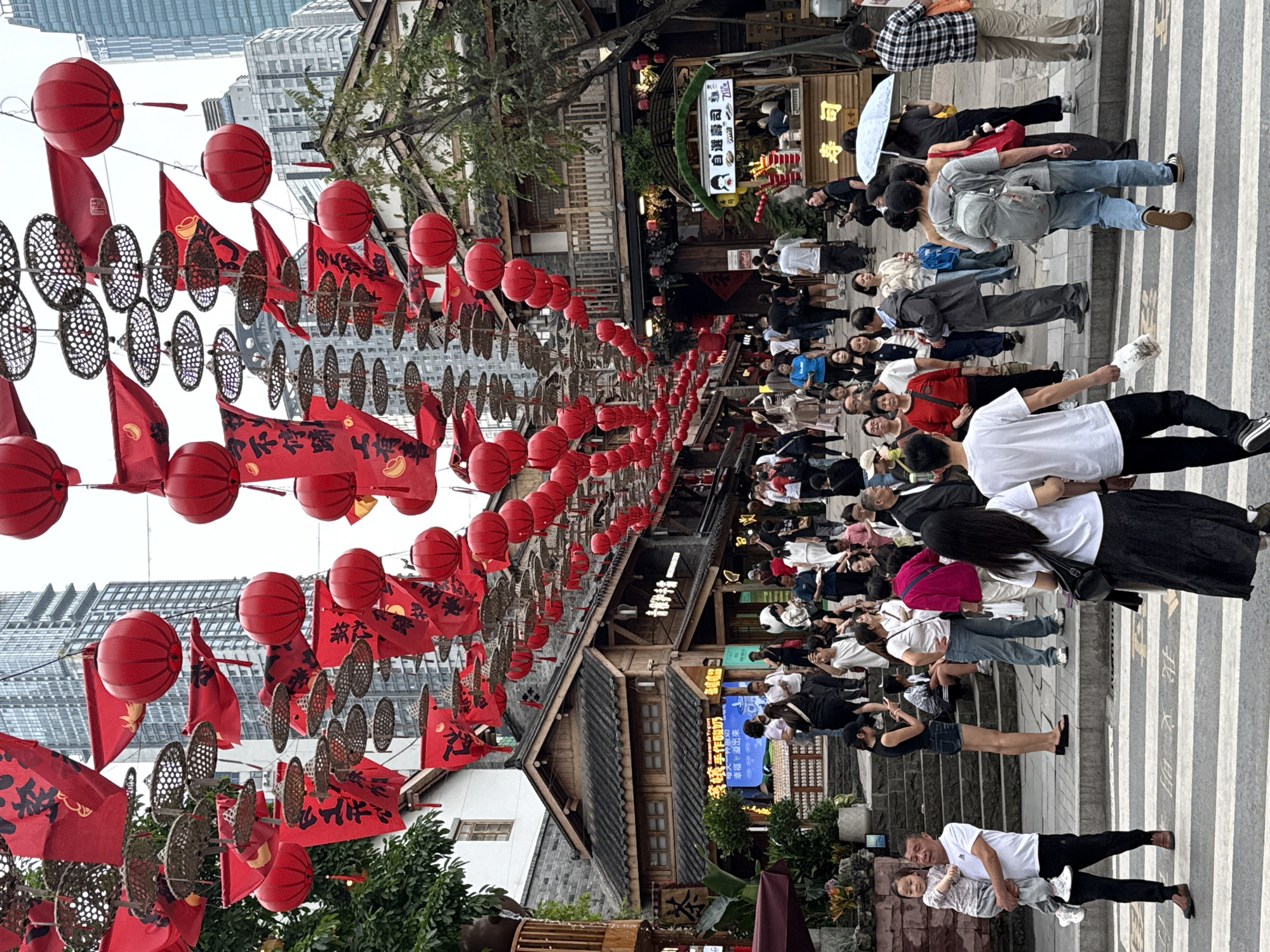 This is a photograph taken in the Yuzhong District of Chongqing, China, and shows a crowded street. Overhead, numerous red lanterns, flags, and circular woven decorations are strung across the street.
A large group of people, adults and children, are walking on the crosswalk. Some are looking at their phones and others are talking amongst themselves. The buildings along the street appear to be traditional Chinese architecture.
On the right of the crosswalk and at the base of the photo, an adult carries a child.
The buildings in the background are tall, modern city skyscrapers. Several food stalls are lined up along the street. There are signs with Chinese writing in yellow and blue color.