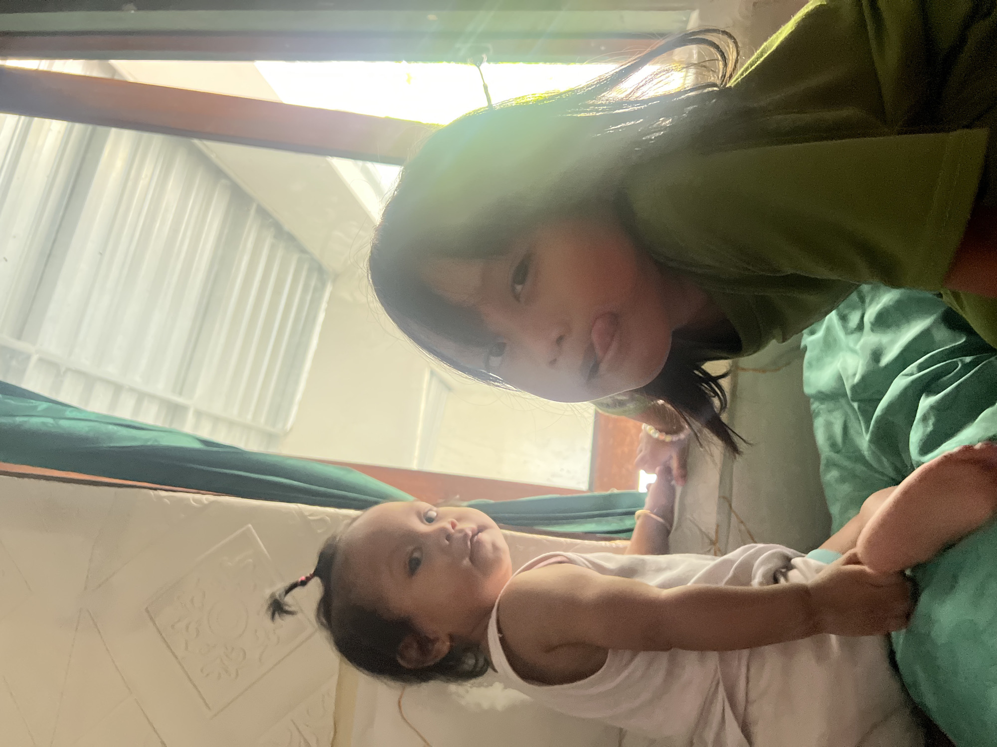 Two young children are seated on a teal-colored blanket or bedspread. The child on the left, facing away from the camera with their head turned to the left, is wearing a white tank top and appears to have dark hair pulled into a small ponytail on top of their head. This child is barefoot and their legs are bent. The child on the right is facing the camera and sticking their tongue out. This child is wearing a green t-shirt and has dark hair. The background includes a window with light streaming in, a green curtain, and a textured ceiling. The setting appears to be indoors.