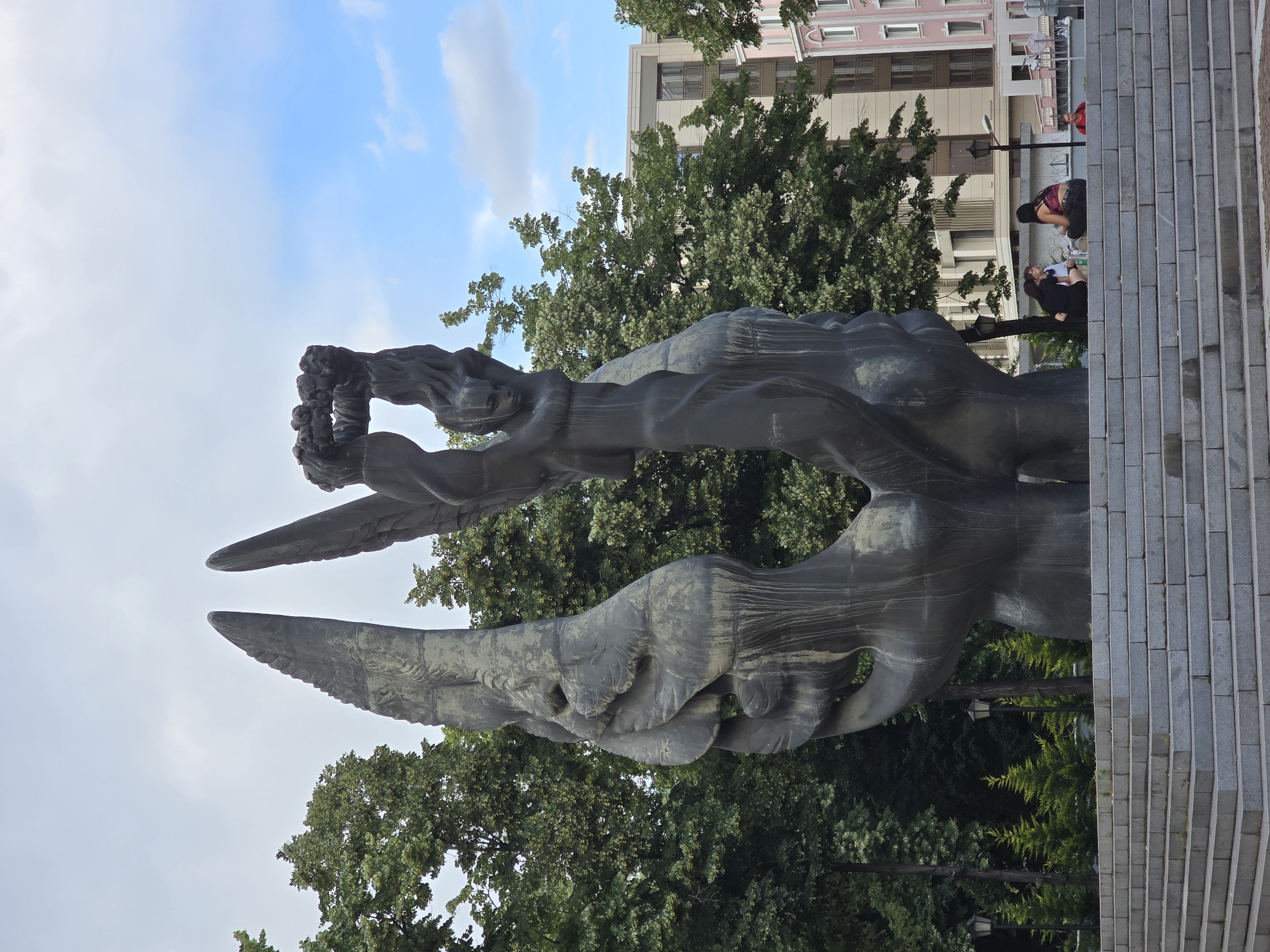 A monumental bronze sculpture depicting two large hands forming a gateway with a wreath of leaves is positioned in front of lush green trees. The sculpture is situated on a wide, tiered granite staircase. In the background, modern buildings with balconies and windows are visible, along with streetlights and smaller evergreen trees. Three people are seated on the steps, facing away from the camera, and a fourth person in a red shirt is visible further down the stairs. The sky is partly cloudy with patches of blue. This sculpture is located on Tsentralen Square in Plovdiv, Bulgaria, near the iconic Roman Stadium.