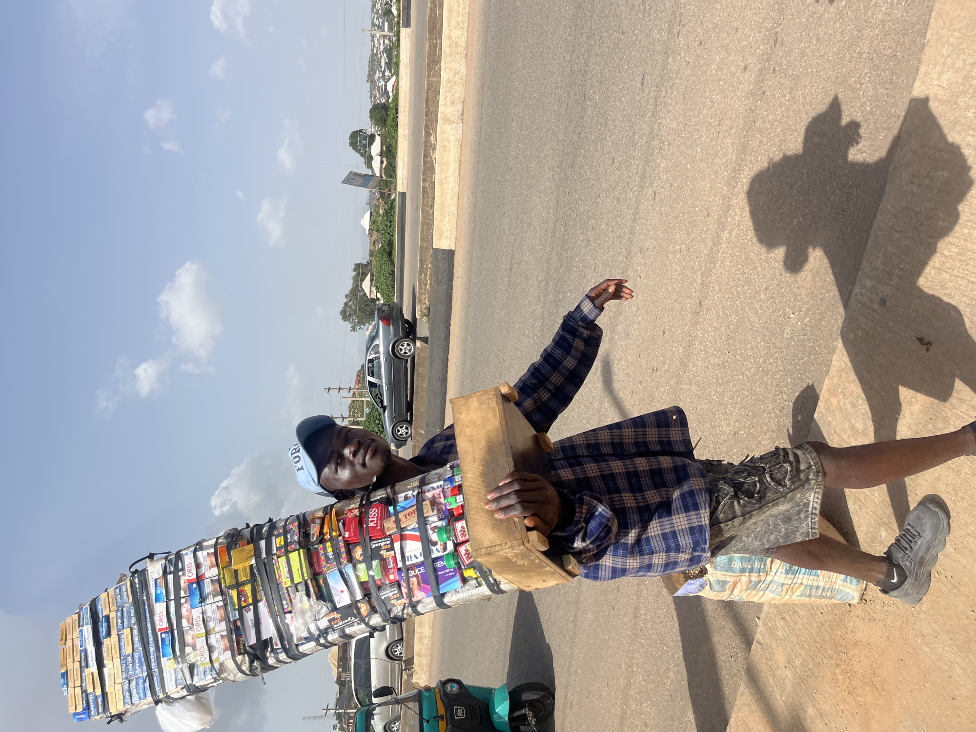 A Black male is walking on a paved road, carrying a large, vertically stacked load of what appears to be cigarette packs and other small items balanced on his head and supported by a wooden structure held in his hands. He is wearing a blue and white plaid long-sleeved shirt, dark shorts, and grey athletic shoes. A light blue baseball cap is on his head. Behind him, to the left, is a turquoise three-wheeled vehicle. To the right, a grey sedan is driving on the road, and beyond that, houses and vegetation are visible on a hill under a partly cloudy sky. The ground is a light brown dirt and asphalt. The location is identified as Gura Top, Nigeria.
