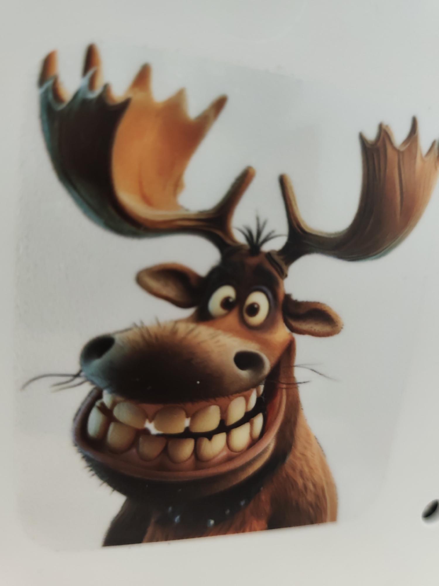 The image presents a close-up, front-facing depiction of a cartoon moose character. The moose features brown fur, large, broad antlers with an orange-brown palette and darker, tined edges, and a dark brown head with a small tuft of messy dark hair positioned between its antlers. Its facial expression includes wide-open eyes with distinct white sclera and dark pupils, a large dark nose from which thin black whiskers protrude laterally, and a very wide, open-mouthed grin showcasing numerous large, yellowish-white teeth across both its upper and lower jaws. A dark collar with small, light-colored studs is visible around its neck. The background is a uniform light greyish-white, suggesting a plain studio setting or a blank surface. A small, dark, circular mark is present in the bottom right corner of the overall photographic capture, indicating the image is a photograph of a physical object. The image itself contains no discernible visual elements or contextual information that would indicate its specific location within Pazardzhik, Bulgaria, or any other geographical area.