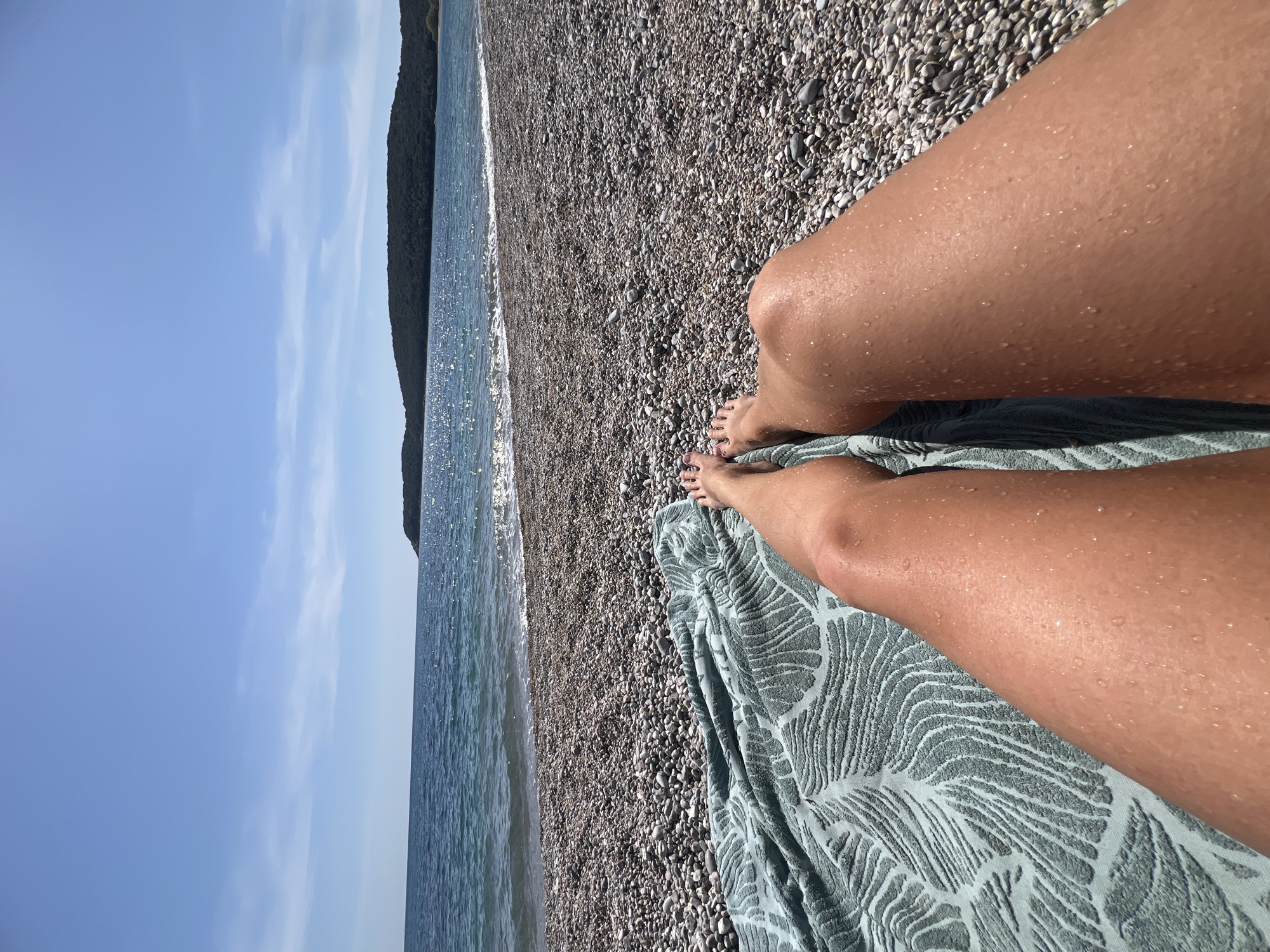 The image shows a person lying on a beach in Prijevor, Montenegro. The person's legs are visible, resting on a patterned blue towel. The beach is composed of small pebbles. The sea is visible, with a coastline and a forested area in the background. The sky is blue with light clouds. The person's legs have water droplets on them, suggesting they have recently been in the sea. The orientation of the image appears to be rotated or taken at an angle.