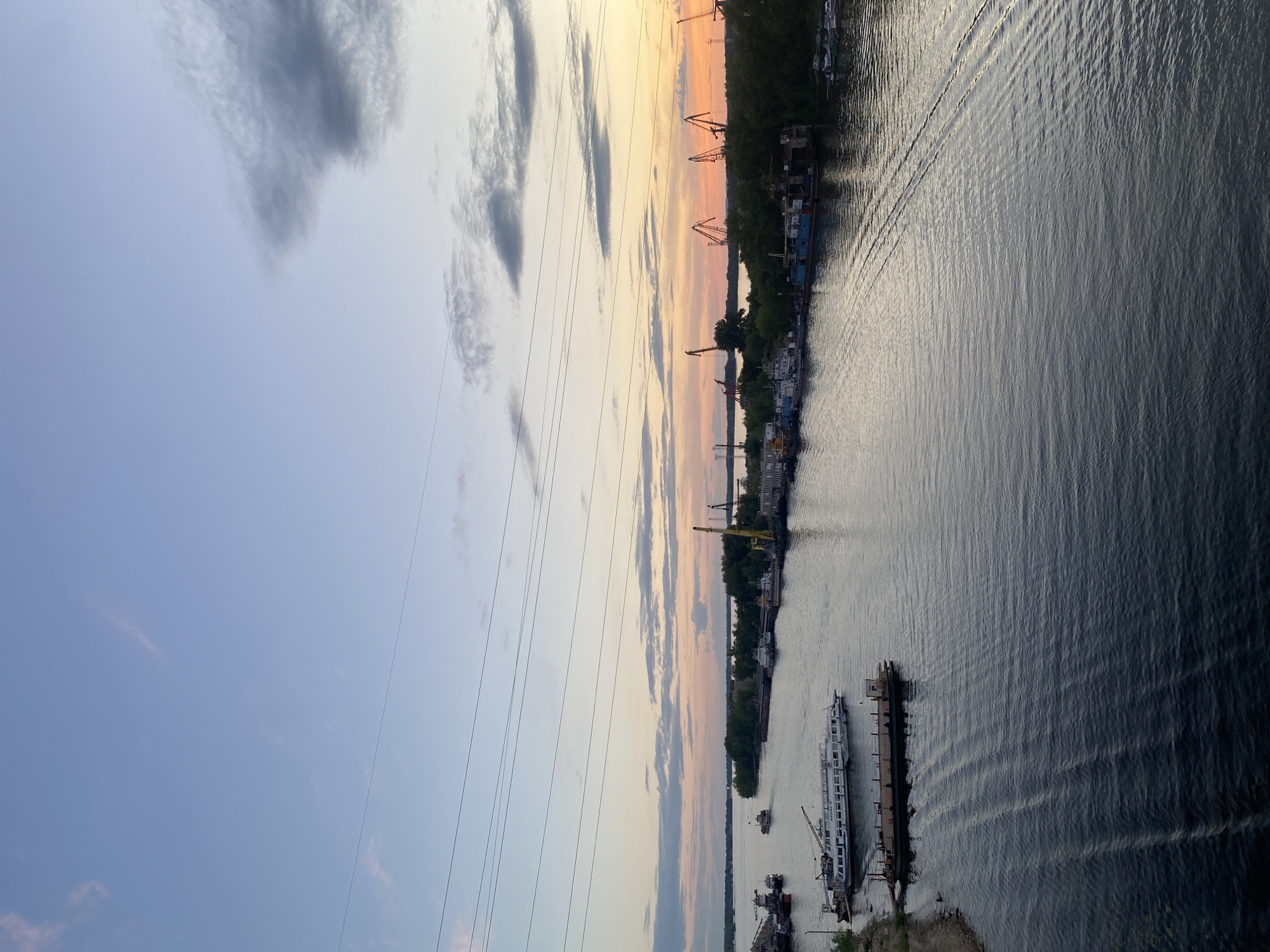 The image depicts a wide river at dusk, with a line of industrial cranes and buildings visible on the far bank, suggesting an industrial port or shipyard. Several boats and barges are moored or navigating the water, with a large passenger vessel and a barge prominently featured in the foreground. The sky is a soft gradient of blue and orange, with wispy clouds, indicating the transition from day to night. Power lines are strung across the upper portion of the frame, adding to the urban or industrial context. The location is identified as Novokuybyshevsk, Russia.