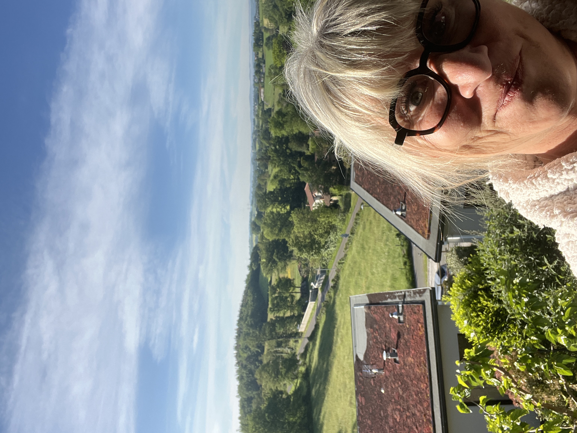 A landscape-oriented, eye-level photograph depicts a woman with gray hair and glasses against a backdrop of Chambœuf, France. The woman's face is visible, positioned in the upper right corner of the frame, and she is wearing eyeglasses. The background includes greenery, rooftops of buildings, a field, and a blue sky with white clouds. The image is bright and appears to have been taken during daylight hours.