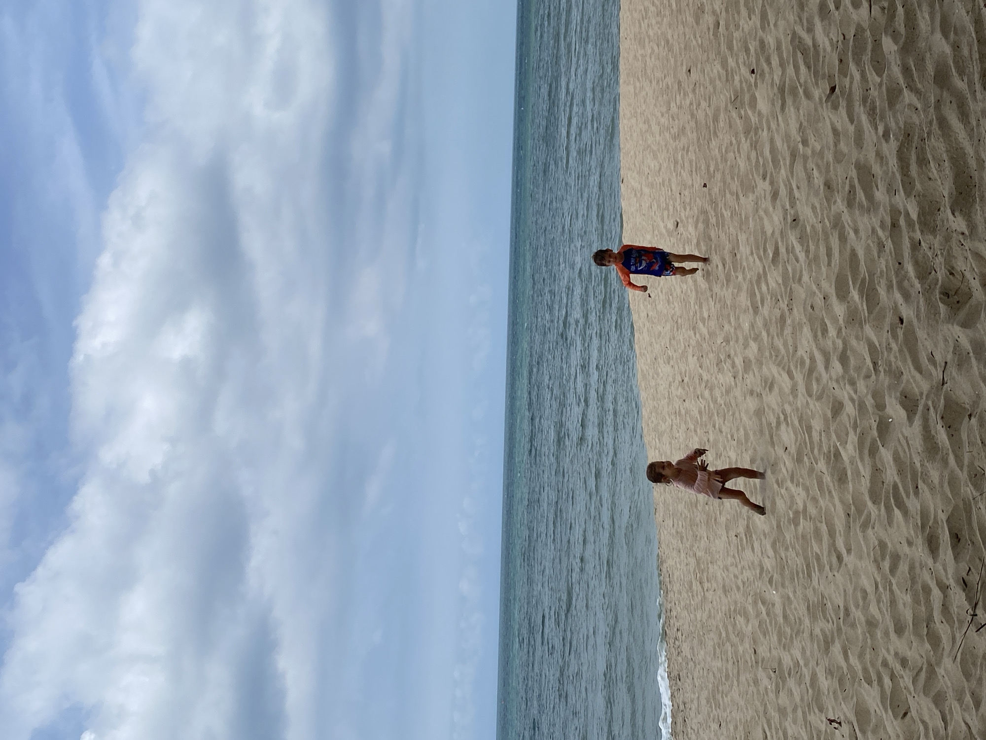 The image depicts two young children on a sandy beach. On the left, a child in a light-colored outfit is partially obscured by a shadow. To the right, another child wearing a long-sleeved rash guard with a blue and orange design and patterned swim trunks is running towards the camera. The children are on a wide expanse of light-brown sand, with gentle waves from a turquoise ocean visible in the background. The sky above is overcast with layers of gray and white clouds, suggesting a diffused light. The scene is likely located at a beach in Phu Thuy Ward, Vietnam, given the context provided.