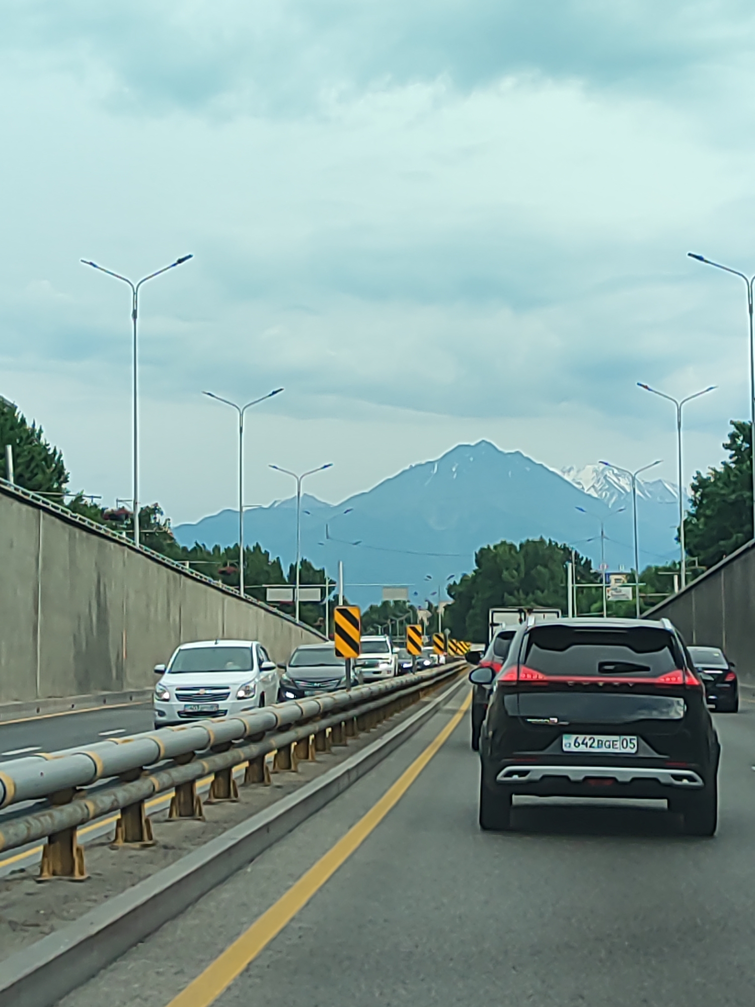 This media file captures a scene of heavy traffic on a highway, likely near Almaty, Kazakhstan. The perspective is from inside a moving vehicle, looking forward. Several cars are visible, all moving in the same direction. The foreground is dominated by a gray concrete barrier with a rusty metal railing, separating lanes. A yellow lane marking runs along the asphalt road.
In the middle ground, a white Chevrolet sedan is prominent, with other vehicles in front and behind it. Further back, a black SUV with a distinctive red taillight design is clearly visible. The license plate on this SUV reads "642 QGE 05".
The background features a striking mountain range, with snow-capped peaks piercing through a cloudy sky. The mountains suggest a mountainous terrain surrounding the city. Tall streetlights line the highway, indicating it's a developed urban or suburban road. The overall weather appears overcast, contributing to a muted color palette. The traffic suggests a busy time of day, likely during peak hours. The scene evokes a sense of urban commuting amidst a grand natural backdrop.