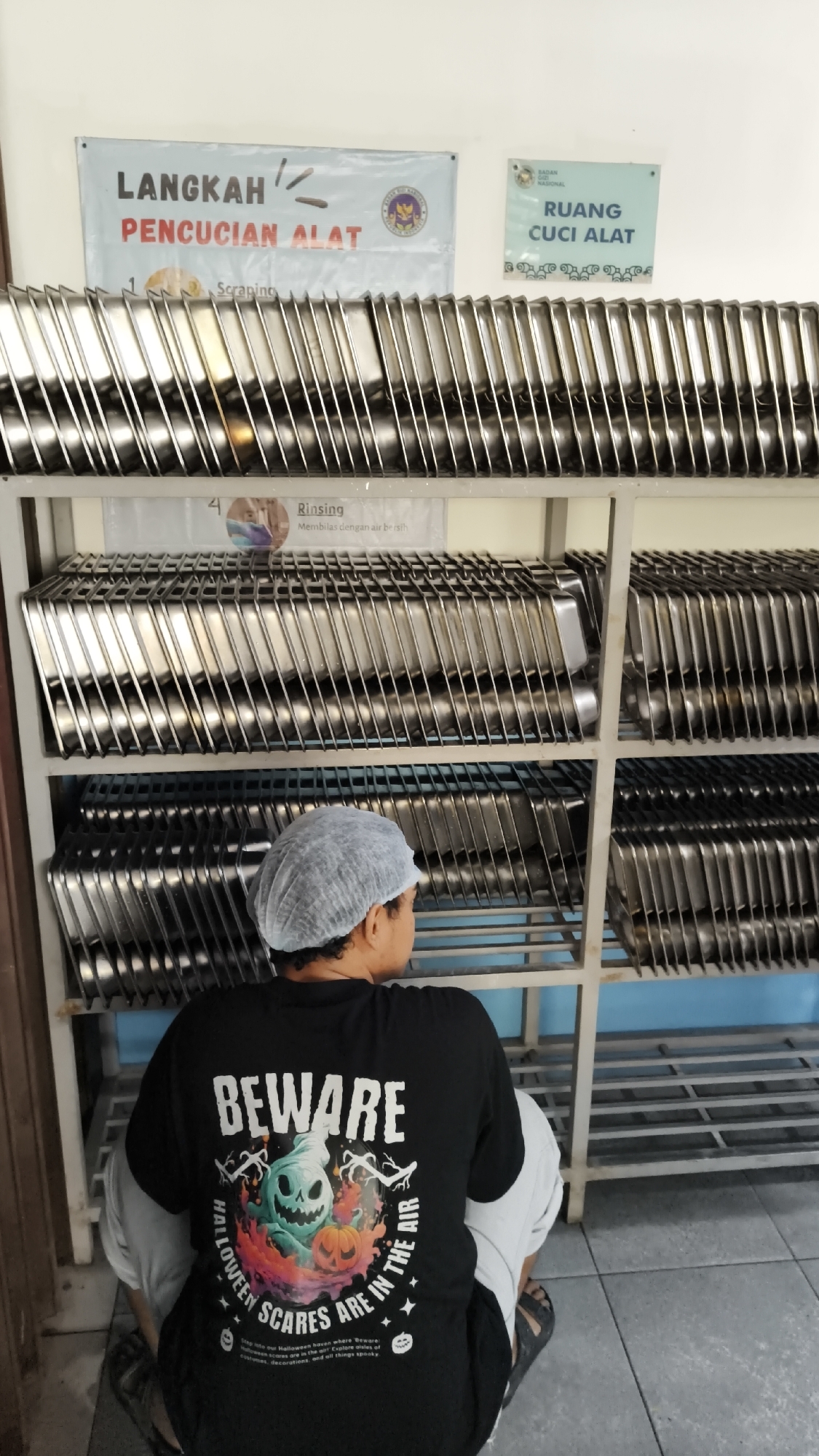 In Sambipondok, Indonesia, a person wearing a hairnet and a black t-shirt with a Halloween-themed graphic is seen sitting on the floor in front of shelves filled with stainless steel trays. The t-shirt's graphic features a ghost, a pumpkin, and the text "BEWARE HALLOWEEN SCARES ARE IN THE AIR." Above the shelves, there are two signs. The left sign reads "LANGKAH PENCUCIAN ALAT" (Steps for Washing Tools) with text that includes "Scraping" and "Rinsing." The right sign is blue and reads "RUANG CUCI ALAT" (Tool Washing Room), indicating the location is a tool washing facility. The environment appears to be an indoor space with tiled flooring and pale walls.