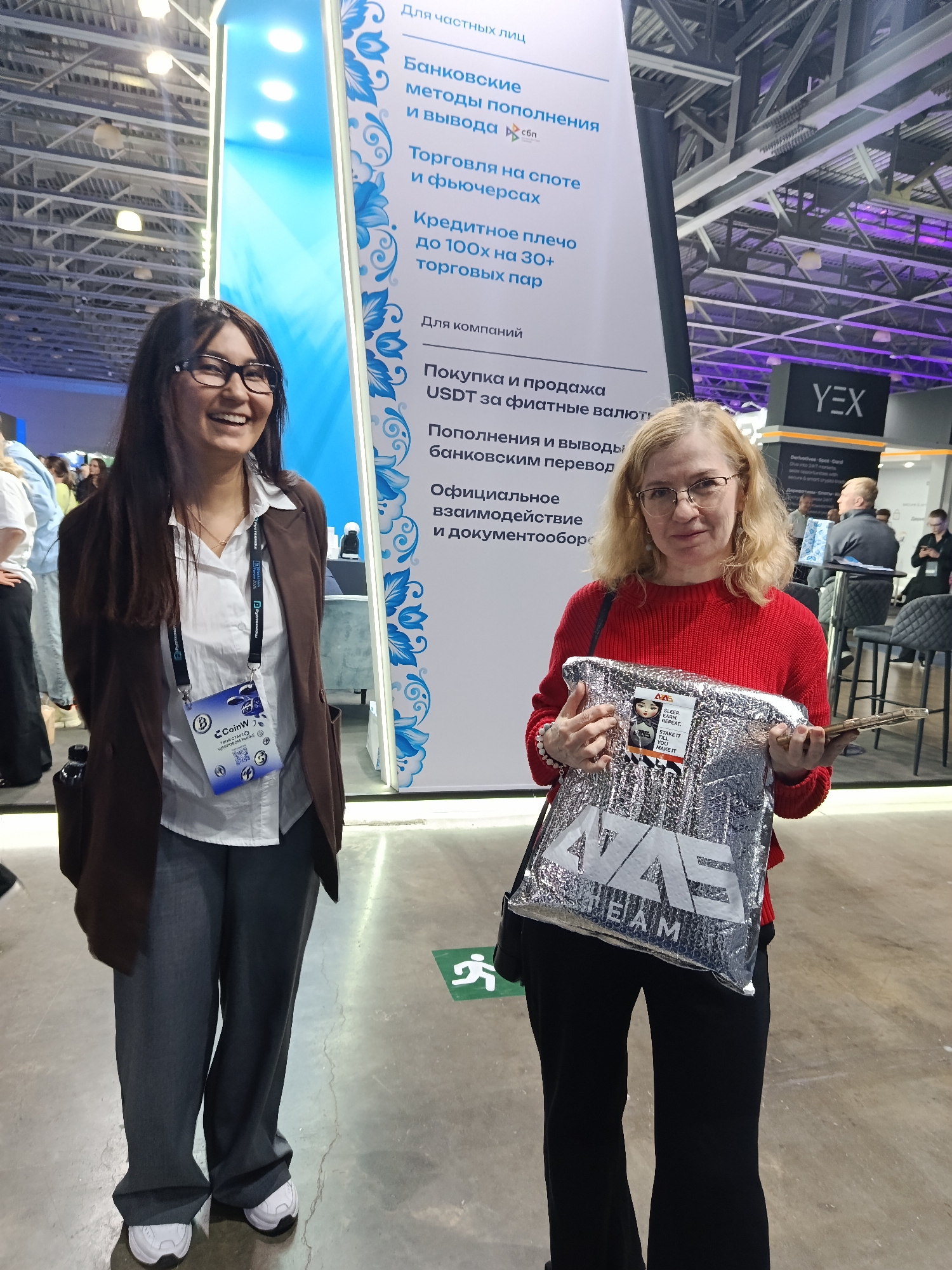 The image shows two women posing indoors, likely at an event or conference in Khimki, Russia.
The woman on the left is wearing glasses, a brown blazer, a white button-down shirt, and grey pants. She has a conference badge around her neck that reads "CoinW". She is smiling and standing next to a sign with Russian text on it.
The woman on the right has short blonde hair and is wearing glasses and a red sweater. She is holding a silver bag with the word "AZAS TEAM" on it. The bag has a sticker on it with the phrase "SLEEP. TRADE. REPEAT. STAKE IT TILL YOU MAKE IT."
In the background, there is another sign with "YEX" printed on it, along with people and chairs. The environment suggests a conference or trade show setting. There is also a green emergency exit sign visible on the floor.
