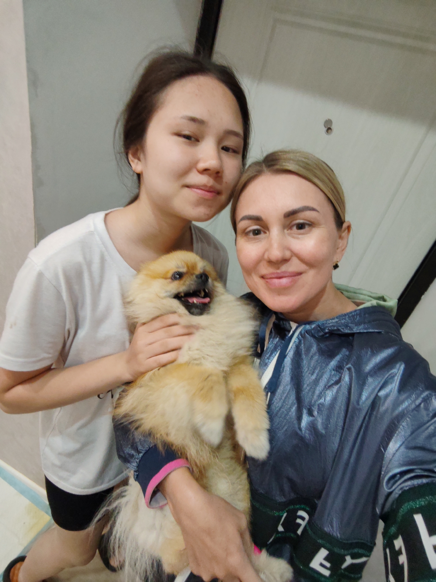 The image shows a selfie of two people, a younger female and an adult female, holding a light brown Pomeranian dog. The younger female is on the left, wearing a white t-shirt and black shorts. She has dark brown hair pulled back and is looking towards the camera with a slight smile. The adult female is on the right, wearing a dark blue iridescent jacket with green trim and white lettering. She has blonde hair and is smiling warmly at the camera, holding the dog in her arms. The dog is fluffy and has its tongue sticking out, appearing happy. The background is an indoor setting, with a plain wall and a white door visible. The lighting suggests it could be daytime. The overall mood is casual and happy, as indicated by the smiles of the people and the dog's expression. There are no prominent weather cues or specific location details beyond the indoor setting, but the context provided is Komsomolsk-on-Amur, Russia.