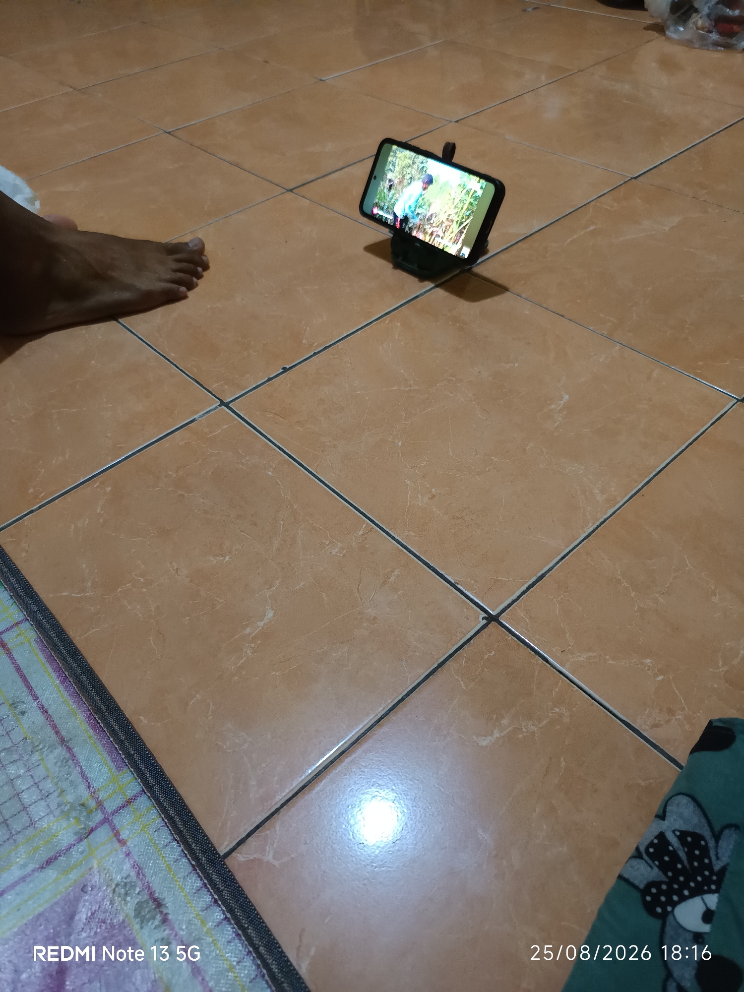 A smartphone is propped up on a black stand on a tiled floor. The phone screen displays an image of a person in a blue shirt in a green outdoor setting. To the left of the phone, a bare foot is visible. The floor tiles are a light brown color with darker grout lines. In the bottom left corner of the image, text reads "REDMI Note 13 5G". In the bottom right corner, a date and time stamp reads "25/08/2026 18:16". The scene is indoors, likely a residential setting.