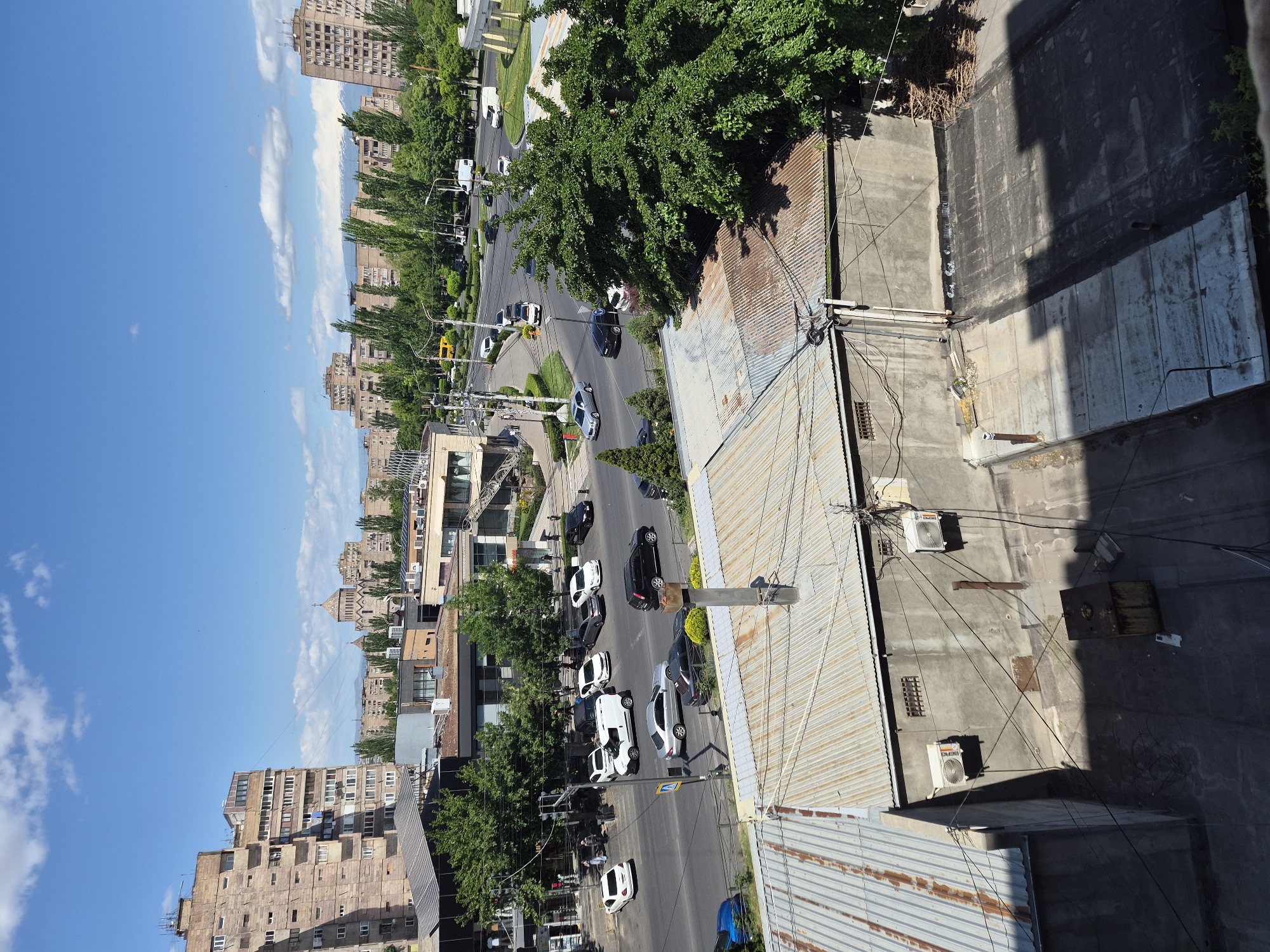 This image presents an elevated view of a bustling urban street in Yerevan, Armenia, captured on a bright, sunny day. The perspective looks down onto the street, which is lined with a mix of multi-story buildings and mature green trees.
The scene features a two-lane road with several cars driving and parked along its sides. Vehicles of various colors, including black, white, and silver, are visible. Lush green trees create a natural canopy over parts of the street and sidewalks. In the background, numerous apartment buildings rise, some appearing older with stone facades, and one distinctive structure featuring a domed roof and intricate detailing, likely a church or historical building. The distant horizon, particularly visible in one of the cropped hints, shows a faint, snow-capped peak, possibly Mount Ararat, under the clear blue sky.
The foreground is dominated by the roofs of adjacent buildings, featuring corrugated metal and flat concrete surfaces. These roofs are cluttered with various elements like utility wires, pipes, and air conditioning units. Shadows are distinctly cast on these surfaces, indicating bright daylight, likely mid-morning or early afternoon. A clear blue sky with scattered white clouds occupies the upper portion of the frame, suggesting fair weather.
Daily urban activity is depicted, with vehicles in motion and parked, and faint figures of pedestrians on the sidewalks. Visible text includes a blue pedestrian crossing sign with a white figure. Close examination of the cropped images reveals additional text on buildings such as "B&B" and "ՕԲՈՆԴ" (OBOND), and on a street sign, possibly "ԿԱՐԱՄԱՆՈՎ" (KARAMANOV). The overall impression is that of a typical, lively city scene.