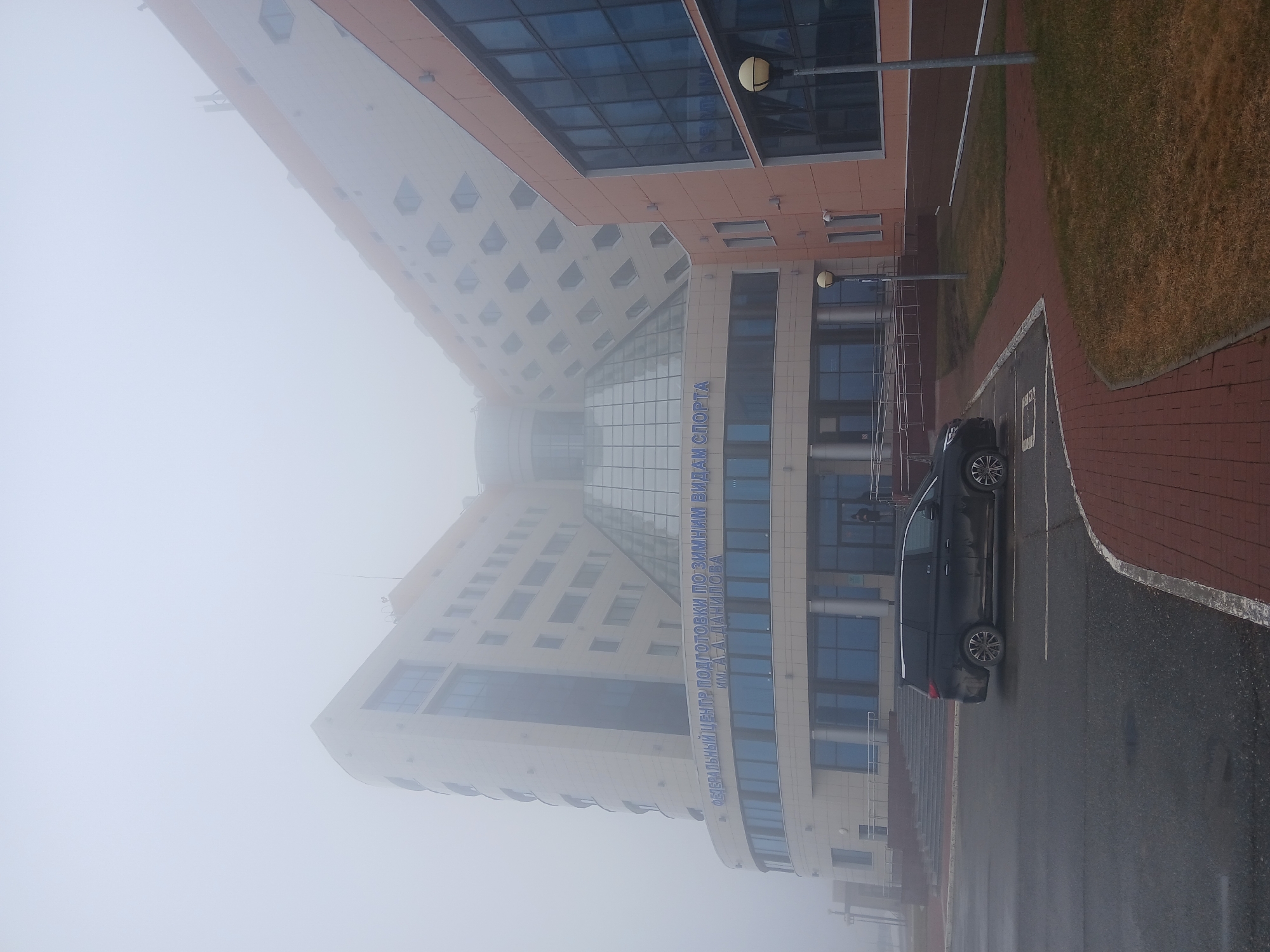 A photograph depicts a building in Прикамский, Russia obscured by heavy fog. The building, identified by blue lettering as the "Federal Center for Winter Sports Training" is a modern structure with a glass facade. A dark-colored SUV is parked in a designated space in front of the building. Brick paving adjoins the parking area, transitioning to a grassy area on the right. Street lamps are visible. The overall scene suggests limited visibility due to weather conditions.