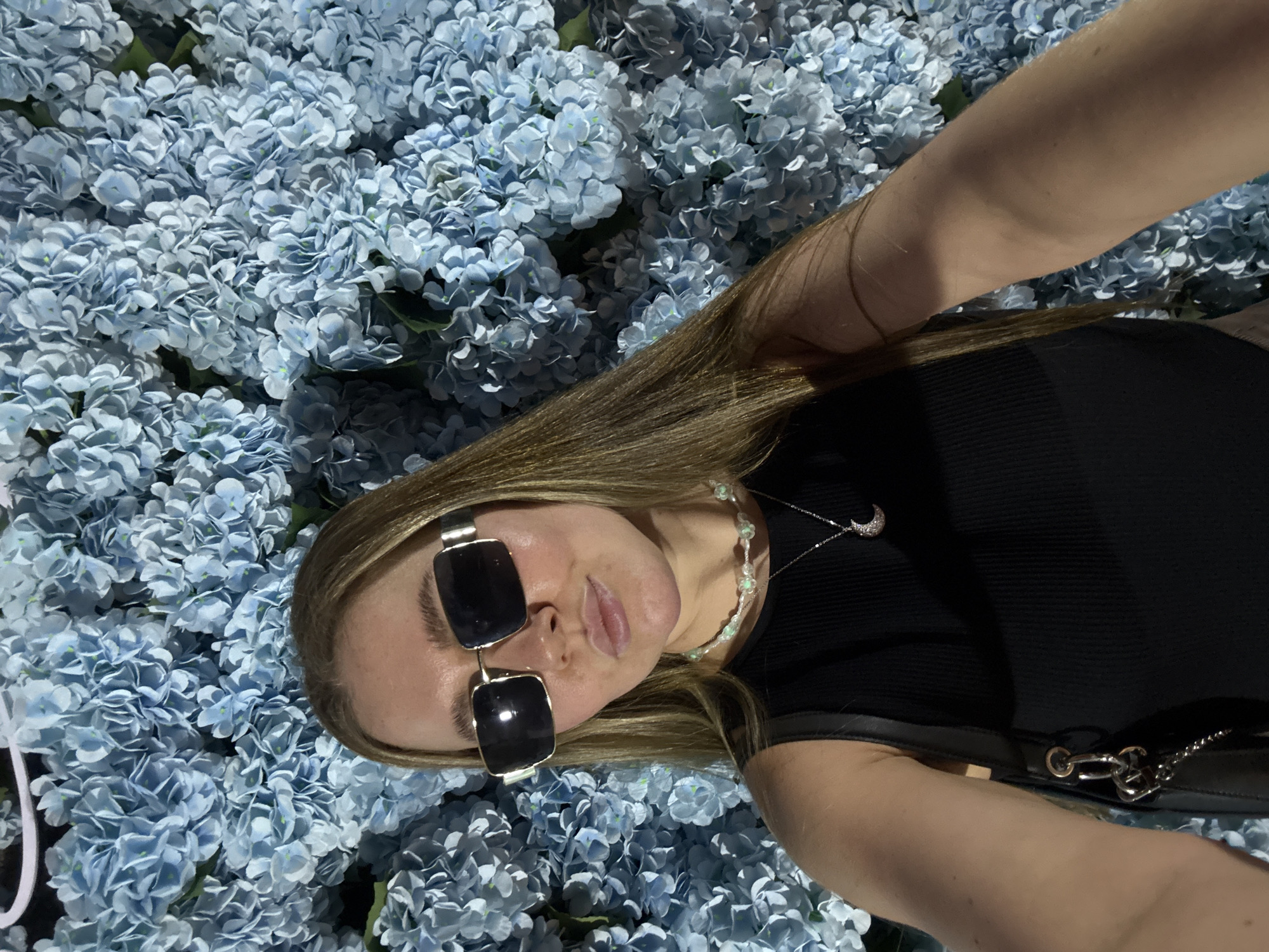 A woman lies on a bed of light blue hydrangeas, with a black ribbed top and black sunglasses. She wears a white beaded necklace and a silver crescent moon necklace. Her hair is light brown. The photograph appears to be a selfie. The city and location are unknown.