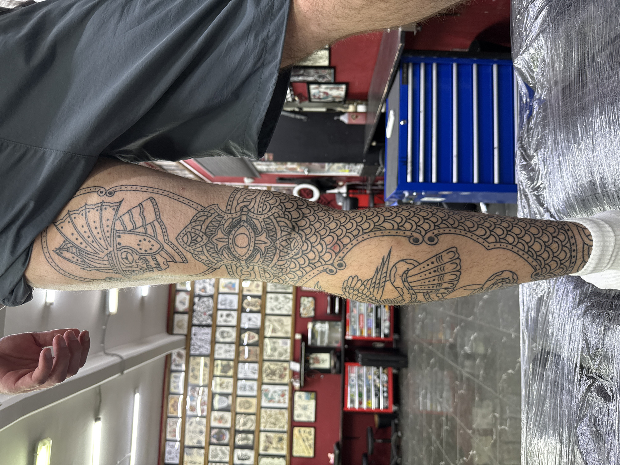 A person is showing a leg tattoo featuring stylized drawings of birds and intricate geometric patterns. The tattoo is on the lower leg, possibly freshly done and covered in clear plastic wrap. The person is wearing gray shorts and a white sock. The background appears to be a tattoo studio, with framed tattoo designs on the walls, a red and black workstation, a blue tool cabinet, and a tile floor. The location appears to be a tattoo studio. The specific city of Moscow, Russia, cannot be confirmed from the image alone.