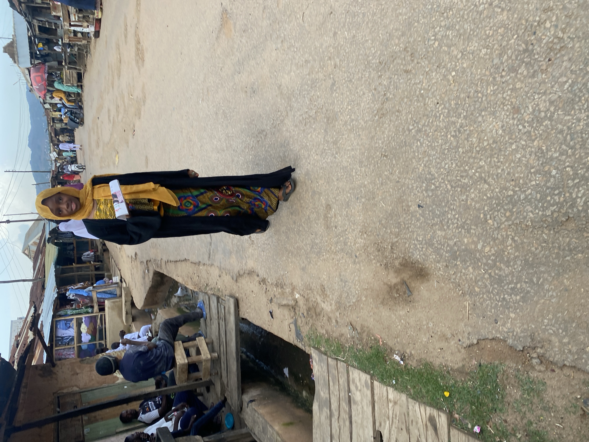 A young Black woman wearing a black abaya and a yellow hijab stands on an asphalt road in what appears to be a market in Jos, Nigeria. She is holding a rolled-up document. In the background, there are several market stalls with corrugated metal roofs, some with goods displayed outside. People are seen walking and sitting near the stalls. To the left of the road, there is a wooden walkway elevated above a dirty, narrow trench or canal. Several men are seated on wooden benches on this side of the road, some of whom are observing the woman. The road is unpaved and has patches of dirt and gravel. In the distance, hills are visible under a cloudy sky. The scene depicts a busy, informal commercial area.