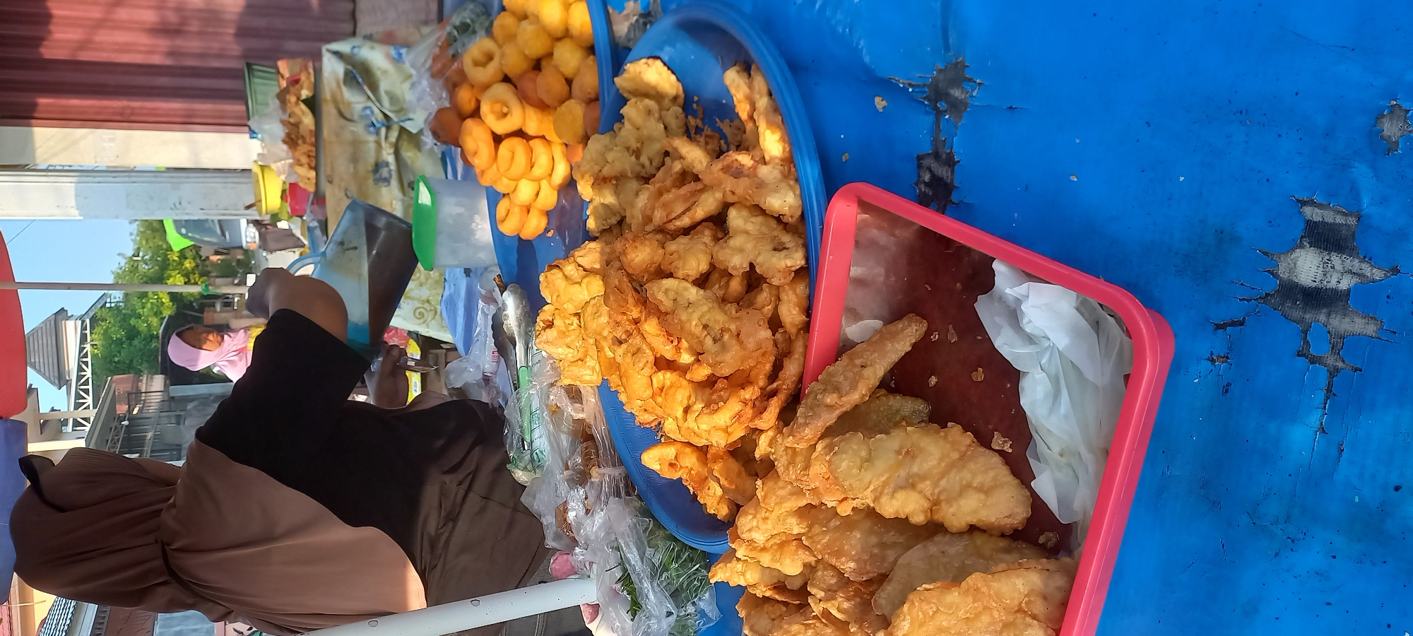 This image depicts a street food stall in Tenggarong, Indonesia. A woman wearing a brown hijab and black shirt is seen from behind, pouring a dark liquid from a transparent pitcher into a container. In the foreground, there are several plates of fried food. A red tray holds pieces of fried food, possibly banana fritters, and a blue tray is filled with more fried items. Behind these, a pile of small, golden-brown, ring-shaped pastries, resembling doughnuts, is visible on another blue tray. In the background, another woman wearing a pink hijab stands near the stall, and a building with a tiled roof is visible. The stall is set up outdoors, with a blue plastic tablecloth covering the surface. The overall scene suggests a bustling market or roadside food vendor.
