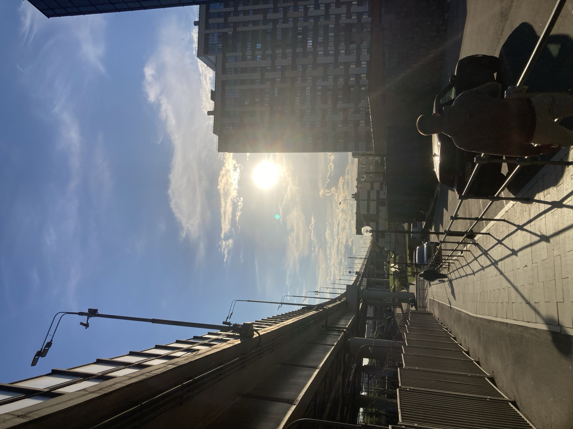 This image captures an urban scene in Moscow, Russia, during either early morning or late afternoon, as suggested by the low-angled sun casting long shadows. The dominant feature is a bright sun, appearing as a clear circle with significant lens flare, against a blue sky adorned with scattered, wispy white clouds.
On the left, a modern, multi-story building with a patterned facade of windows and dark panels rises prominently. An elevated concrete and metal structure, likely a bridge or viaduct, runs diagonally across the middle of the frame from the foreground towards the background, featuring robust railings and supports.
To the right, a paved sidewalk with grid patterns and a darker road are visible. A shirtless man stands next to a dark-colored parked car, his back partially to the viewer, and his figure silhouetted by the sunlight. Long shadows from the man, the car, and railings extend across the pavement. The car's license plate appears to have Cyrillic characters, reinforcing the location context. Streetlights are mounted along the elevated structure and also visible further down the road. The overall scene depicts a typical city environment under bright, direct sunlight.