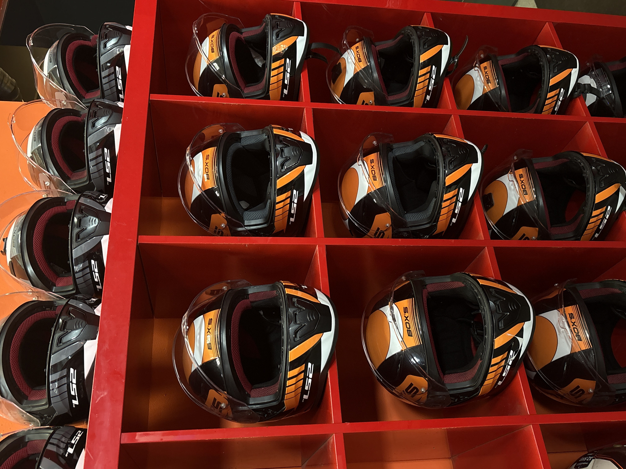 The image shows a retail display of multiple motorcycle helmets, all black with orange and white accents. The helmets are arranged in a red shelving unit with individual compartments. The top row of the shelving unit contains three helmets, each with a clear visor. The second row also contains three helmets, identical in design to the top row. The third row displays two helmets, one of which is partially visible. The bottom row shows two more helmets, with the one on the right also partially obscured. The overall scene suggests a retail environment, likely a motorcycle shop. The image does not contain any people or any contextual information that would help pinpoint its location in Saint Petersburg, Russia.