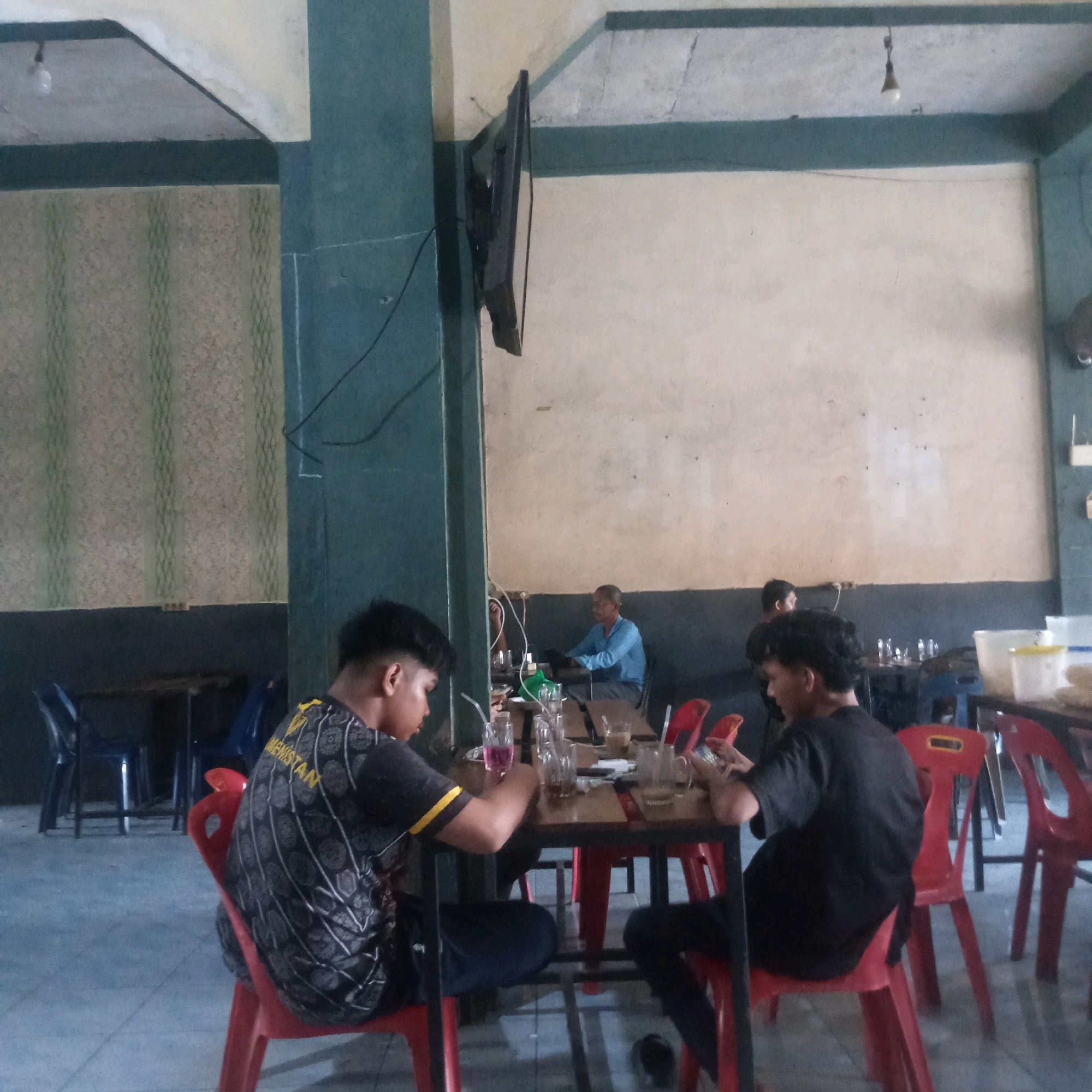 The image depicts the interior of a cafe in Grong-Grong, Indonesia. The cafe features a light-colored, textured wall and dark teal accents on the columns and ceiling trim. A wall-mounted television is visible on the left side. Several tables with chairs are arranged in the space. In the foreground, two young men are seated at a table. The man on the left is wearing a black t-shirt with a yellow and black patterned design and is looking down at his phone. On the table in front of them are several glasses, some with drinks and straws, and a phone. The man on the right is wearing a black t-shirt and also appears to be looking at his phone. Further back, another man in a blue shirt is seated at a table, and a glimpse of another person is visible in the background. Red plastic chairs are a prominent feature around the tables. The floor is tiled.