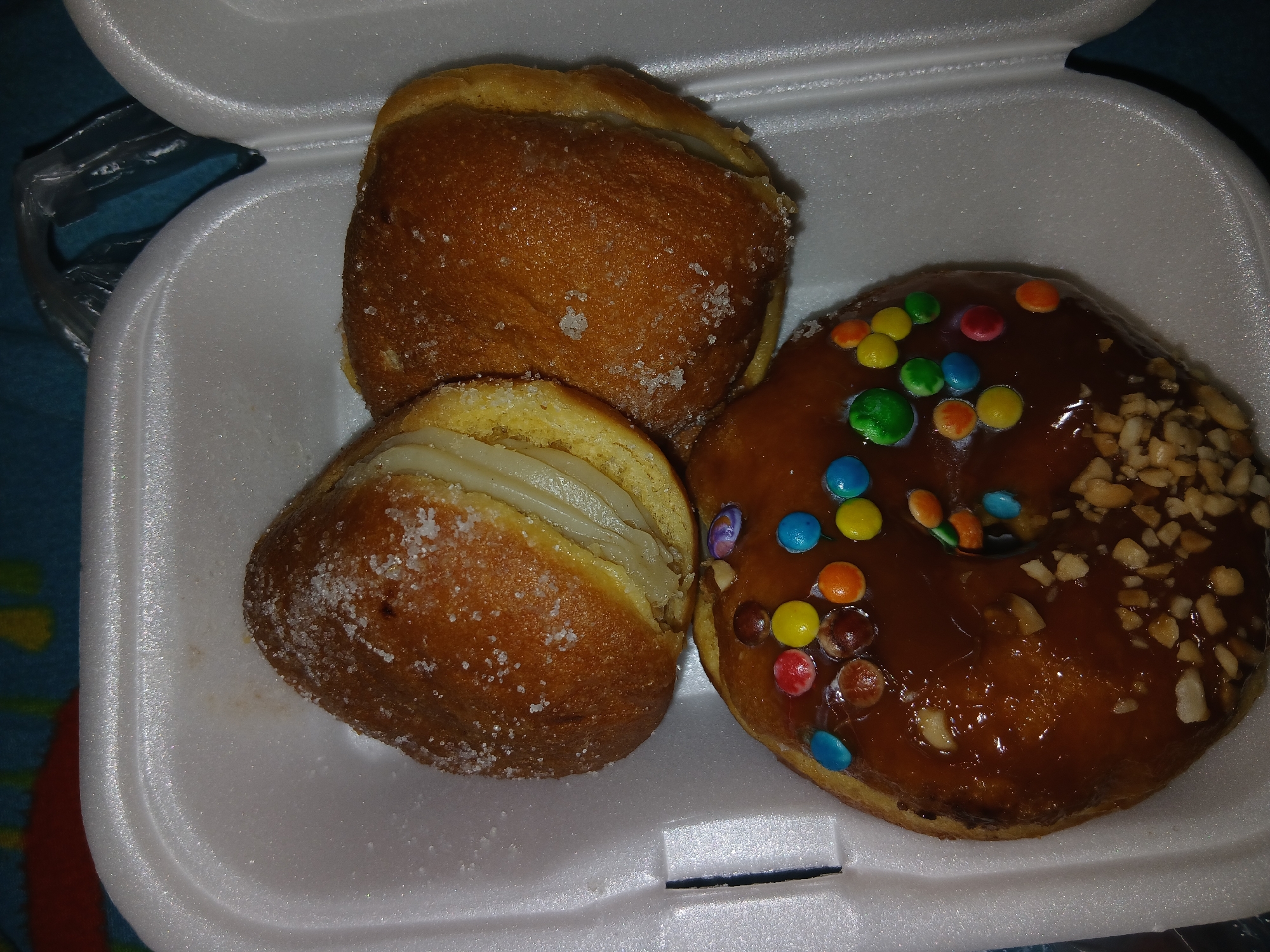 A white styrofoam container holds three donuts. Two donuts are coated in granulated sugar, one sliced and filled with a white cream. The third donut is glazed with brown icing, decorated with colorful candies and chopped nuts. The location is not discernible from the image. No people are visible.